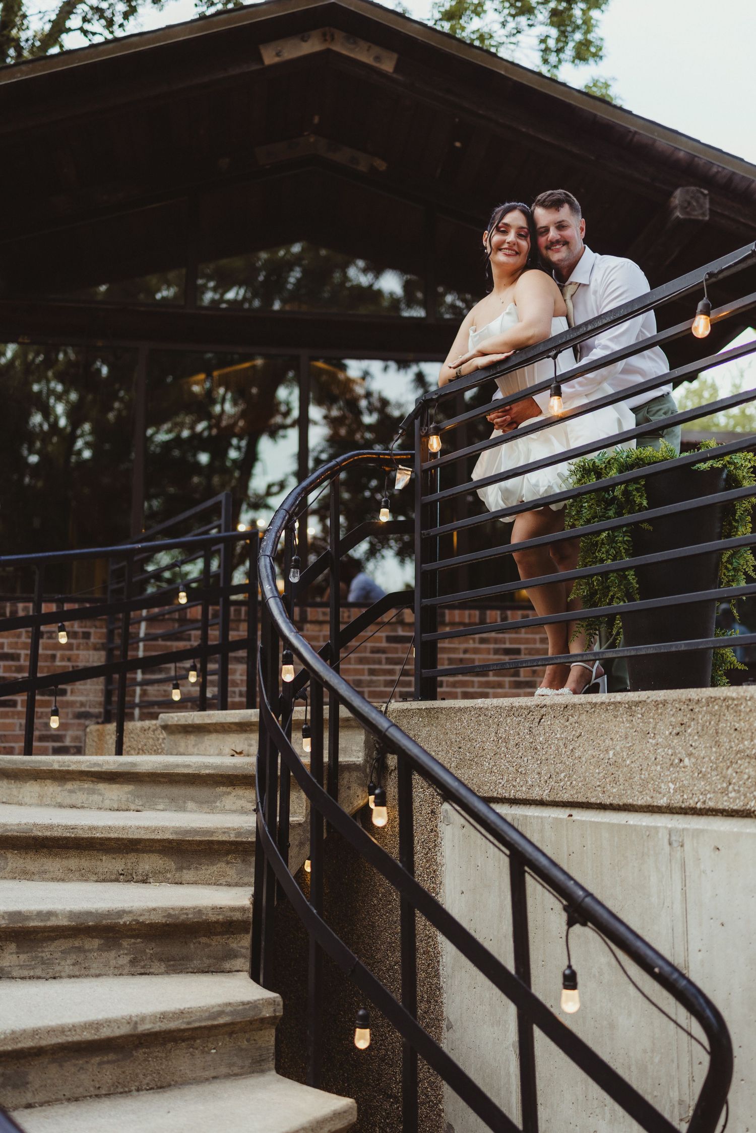 Smiling bride and groom pose on decorated staircase with string lights at dusk.