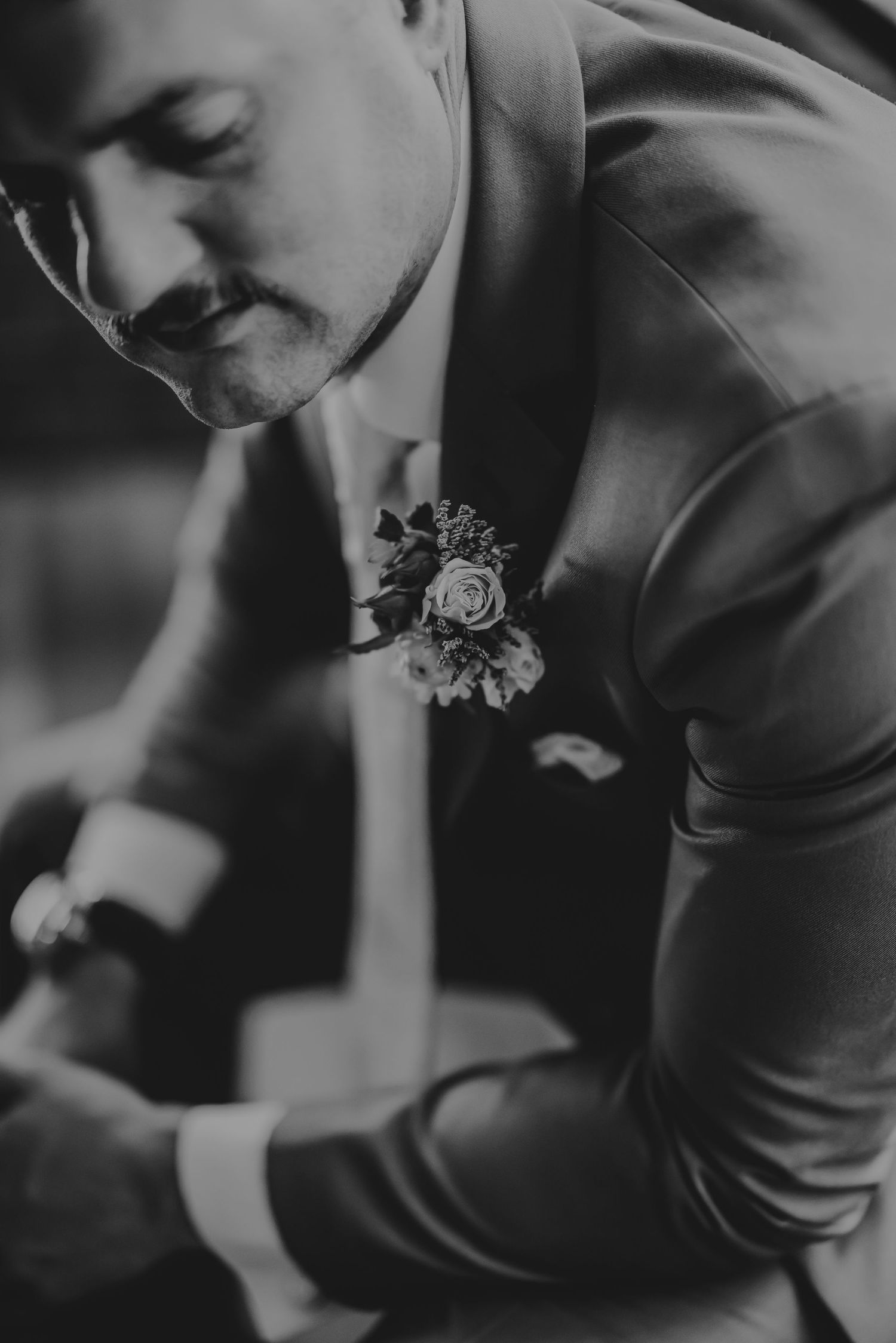 Black and white photo of a groom in a suit with a floral boutonniere, looking down while adjusting his cufflinks.