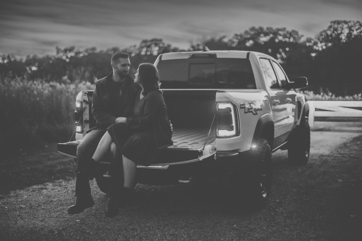 Black and white photo of couple sitting on truck tailgate at sunset.