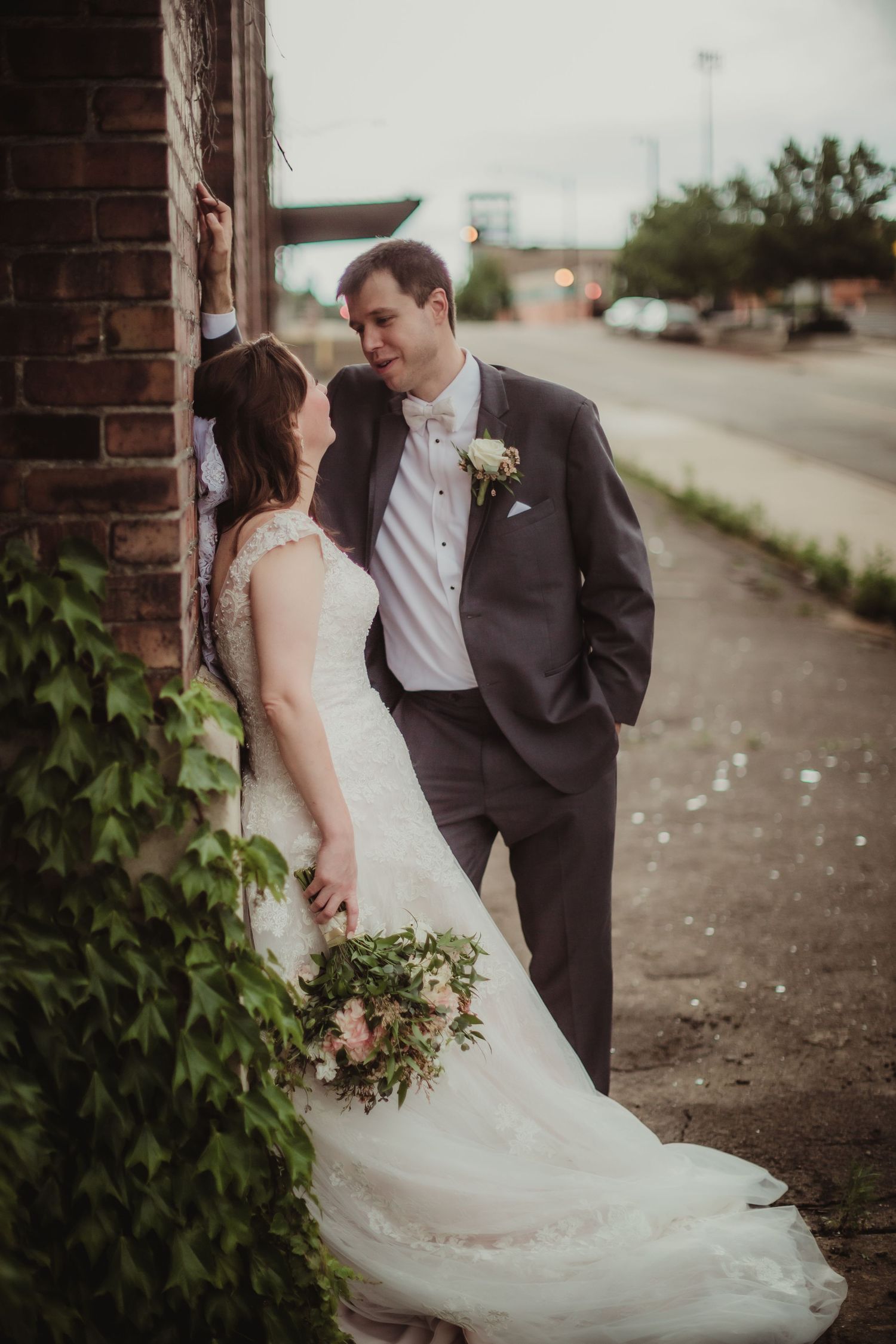 Newlyweds share an intimate moment against a brick wall with greenery, the bride holding a rustic bouquet.