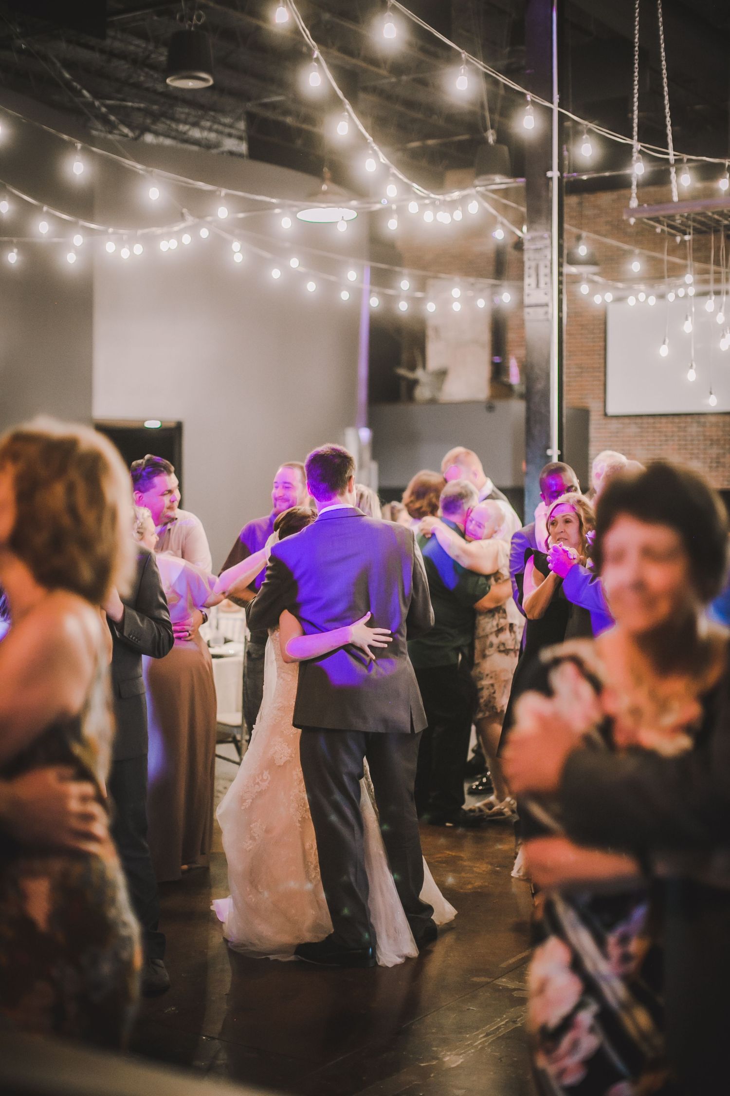 Wedding guests dance under string lights in a modern industrial venue space.