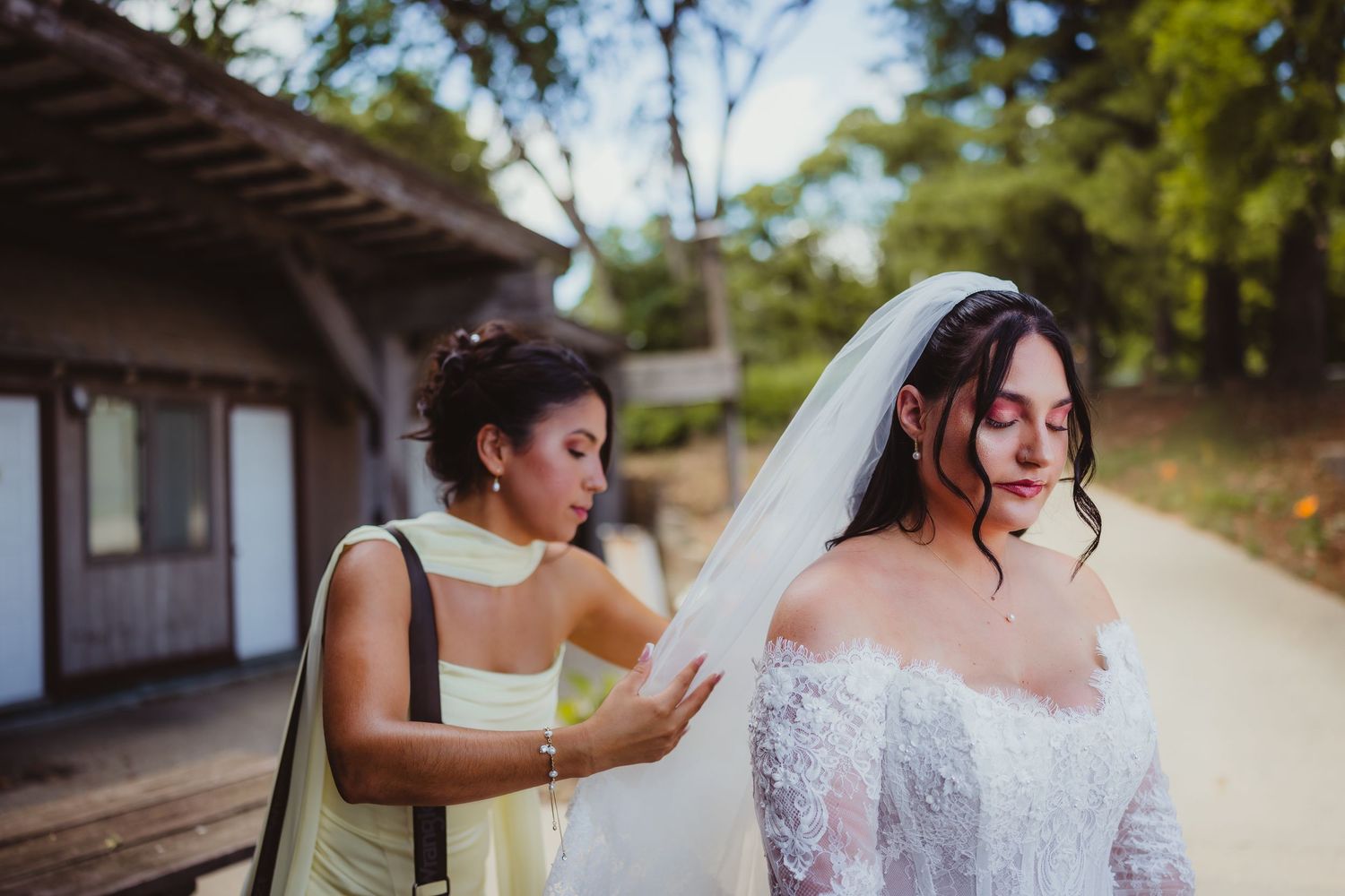 Bridesmaid in yellow dress adjusting bride's white veil outdoors at rustic wedding venue surrounded by trees.