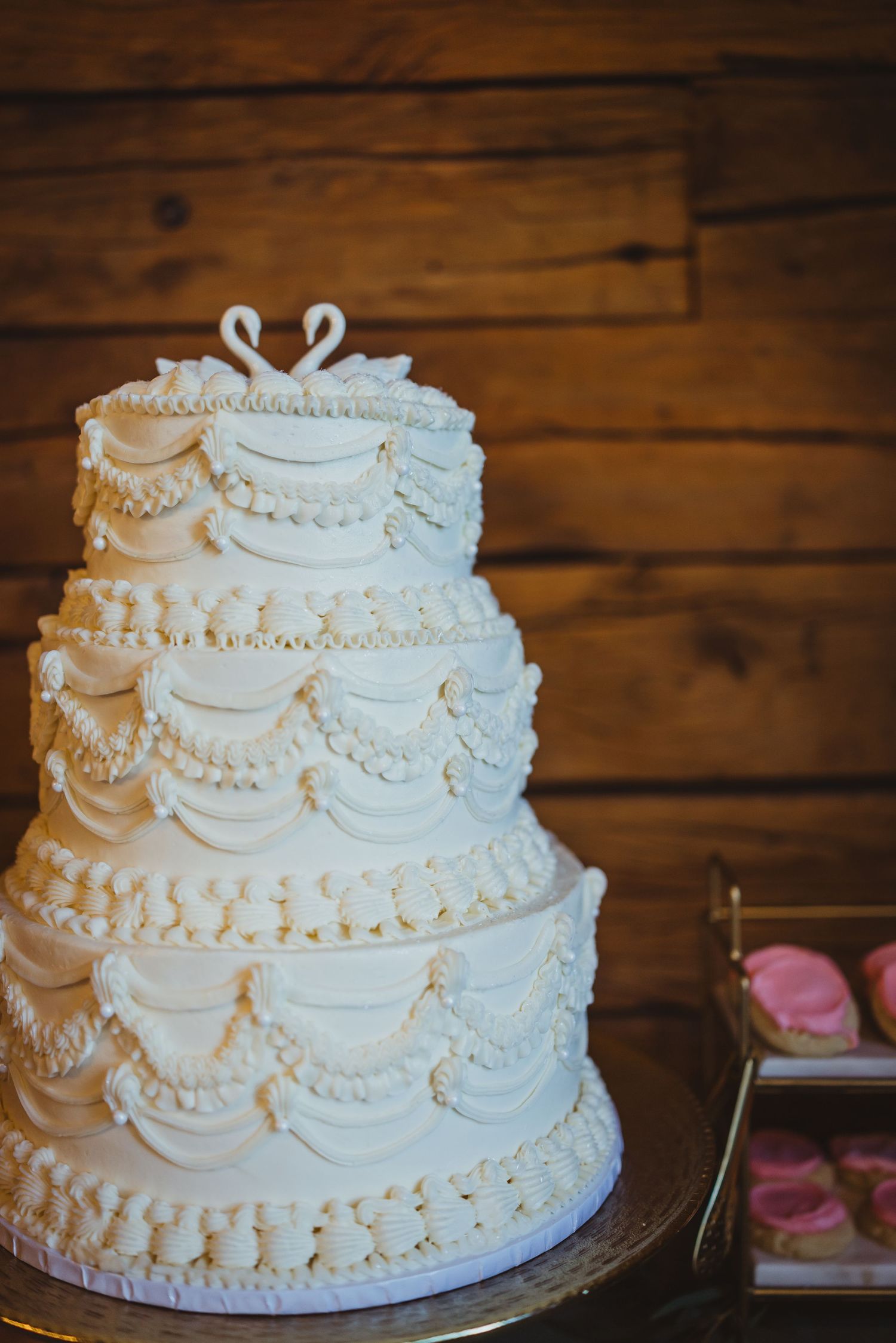 Elegant three-tier white wedding cake with swan topper, ruffled frosting details, and pearl borders on a rustic wood backdrop.
