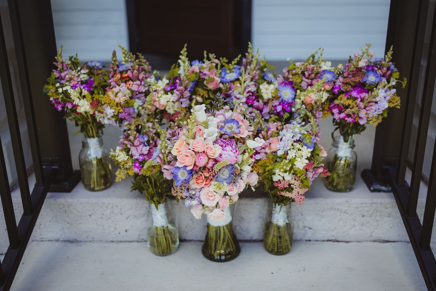 Seven colorful wildflower wedding bouquets in glass vases arranged on white steps for a rustic bridal party.
