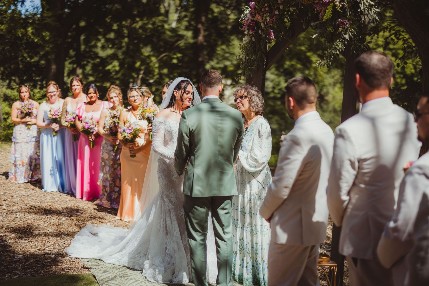 Outdoor wedding ceremony with bride and groom at altar, colorful bridesmaids in mismatched dresses, surrounded by lush green trees.