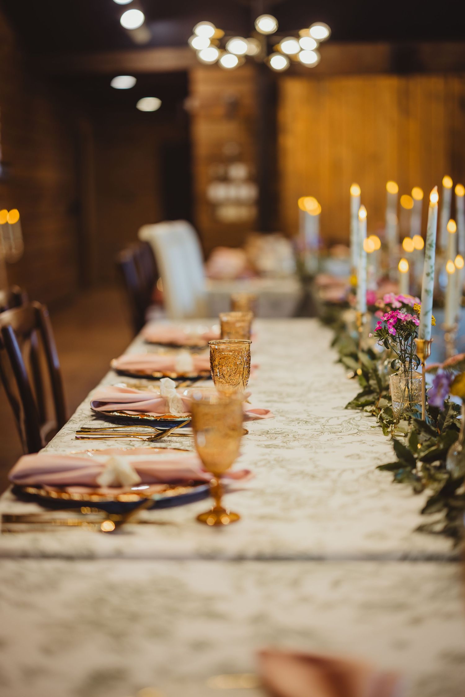 Long elegant wedding reception table with amber goblets, pink napkins, lace runner, and candles in a rustic barn.