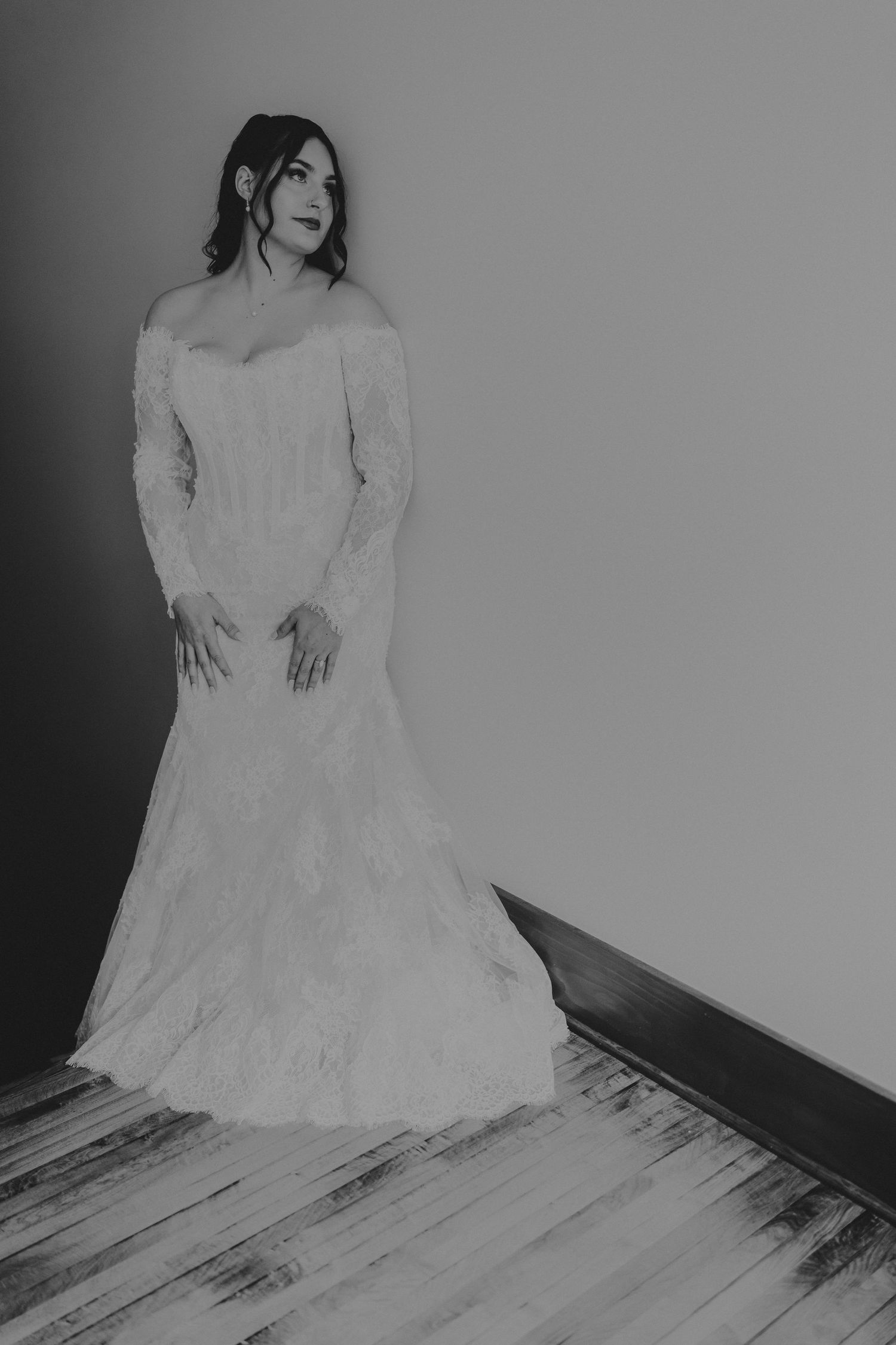 Black and white photo of a bride in an elegant off-shoulder lace wedding gown with long sleeves, posing on a wooden floor.