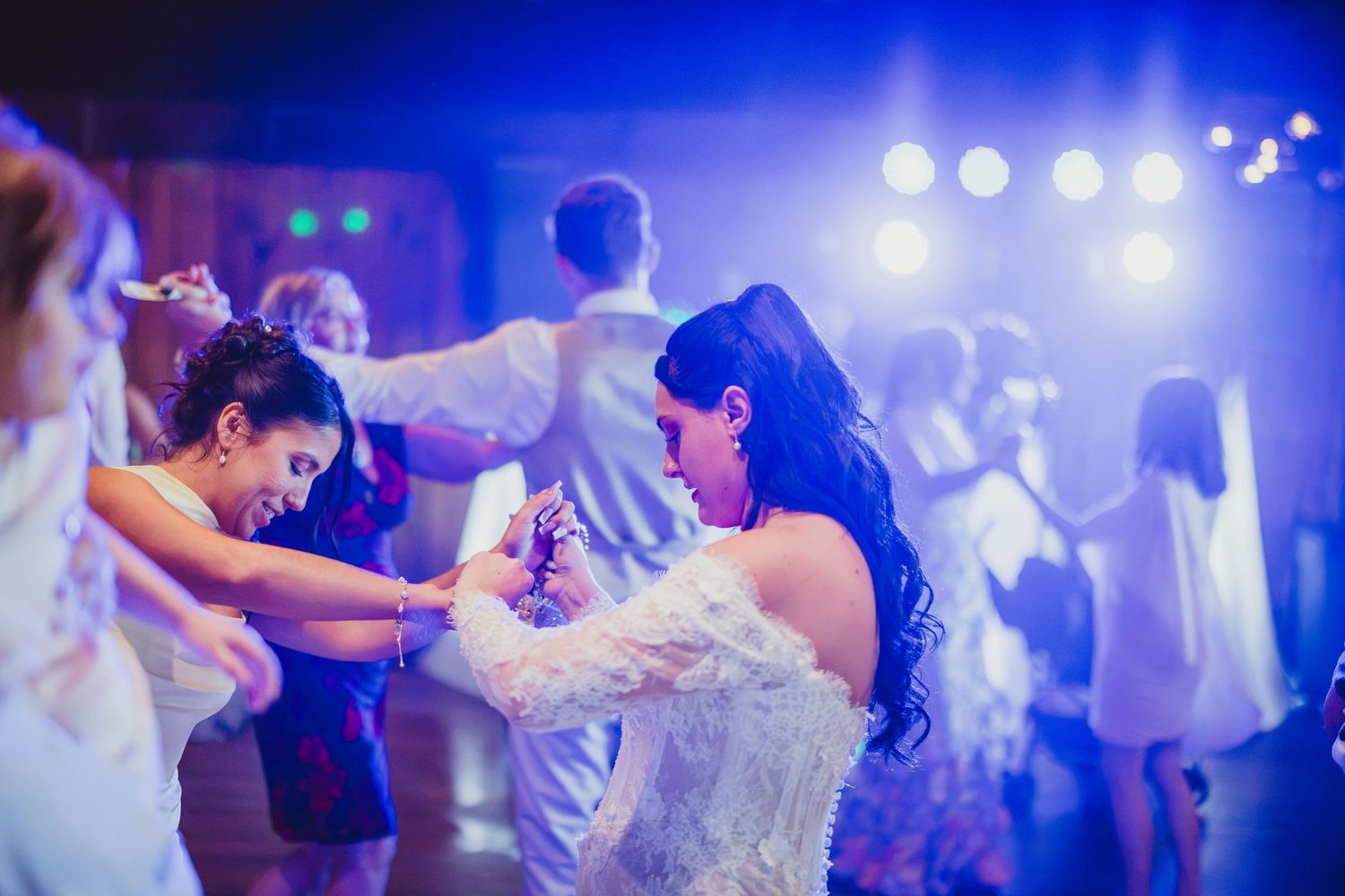 Two women in white dresses dancing joyfully at a lively wedding reception under blue stage lights.