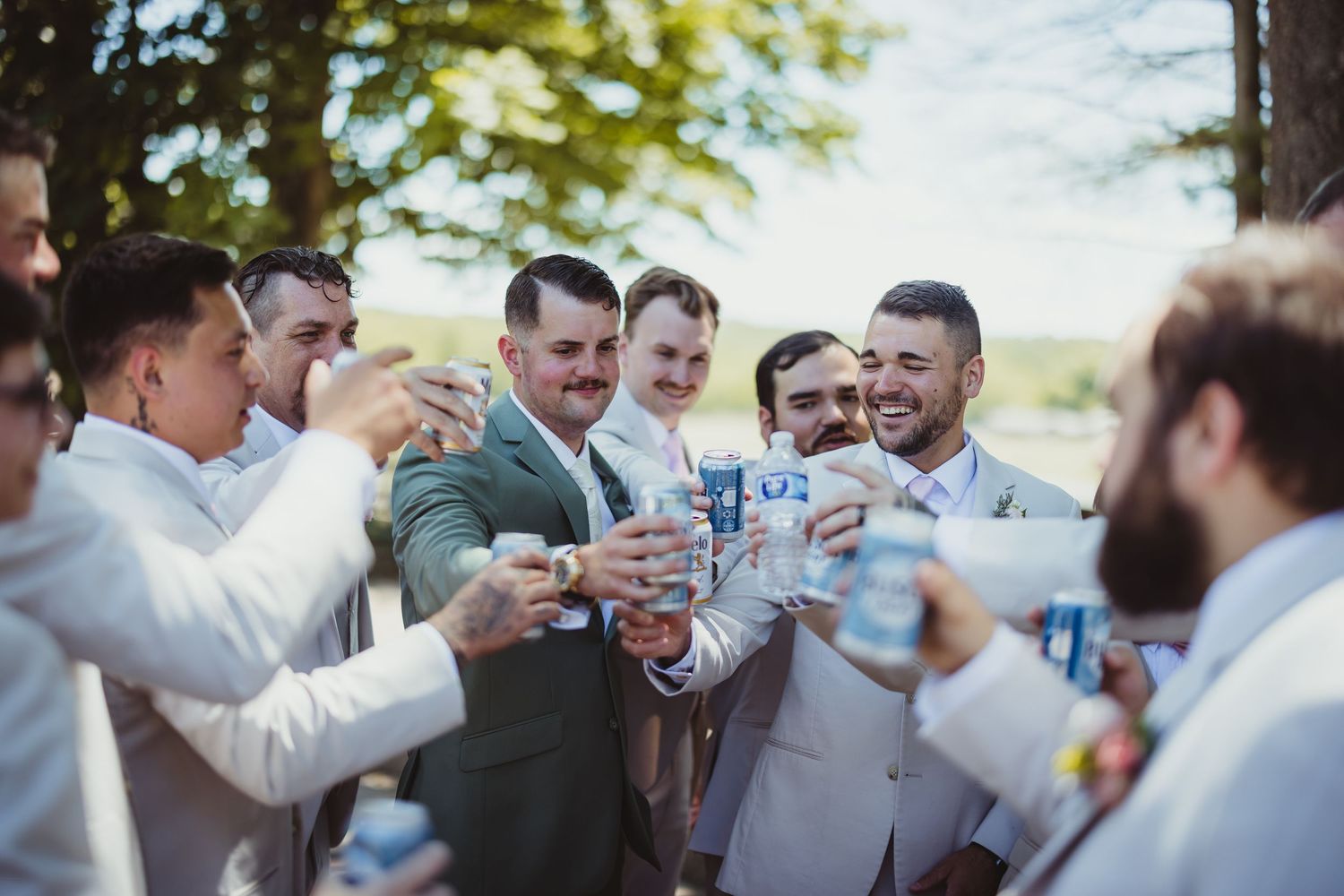 Groomsmen in suits toasting with beer cans outdoors at a wedding celebration.