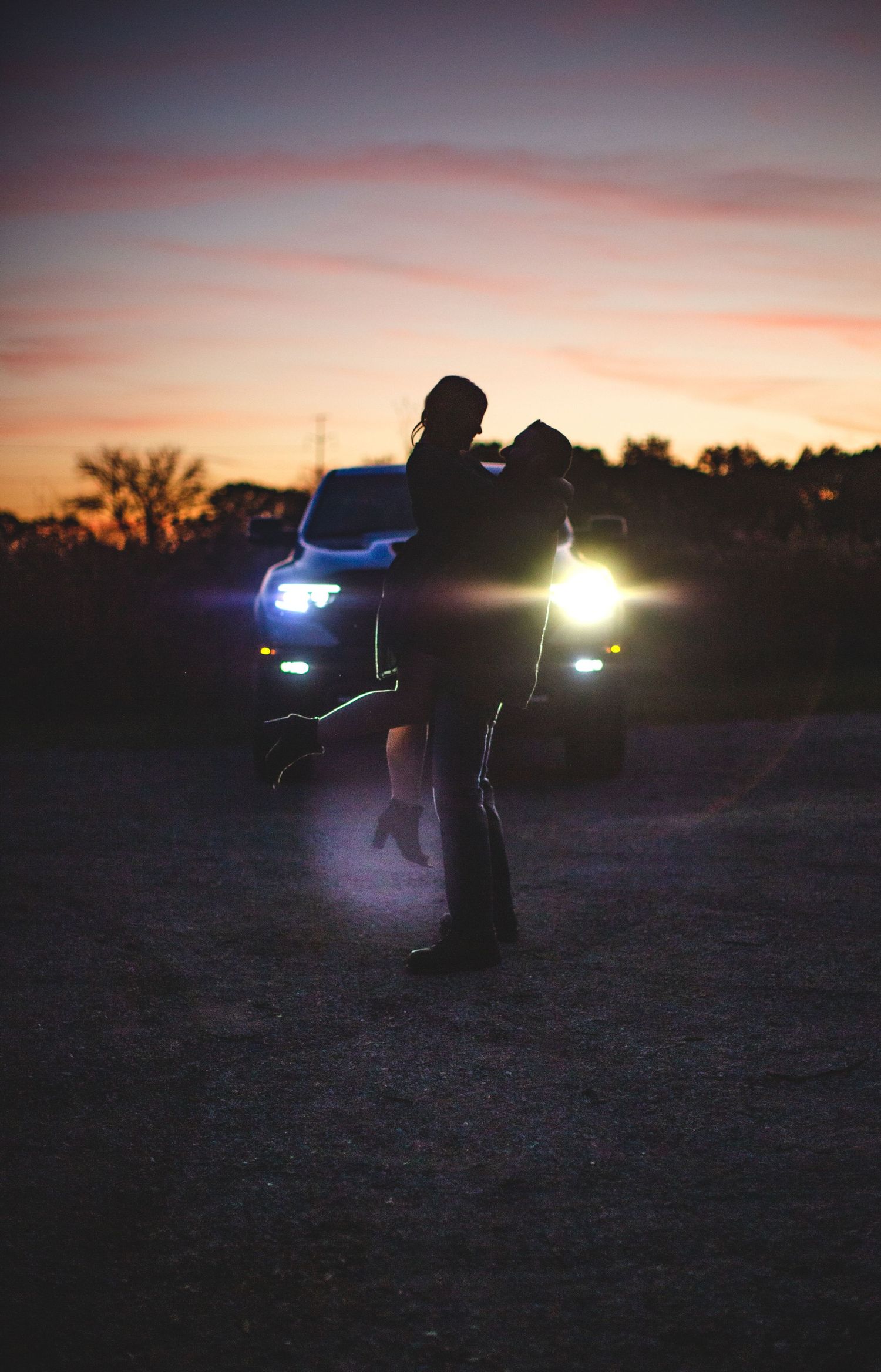 Silhouette of figures embracing near car with headlights at sunset.