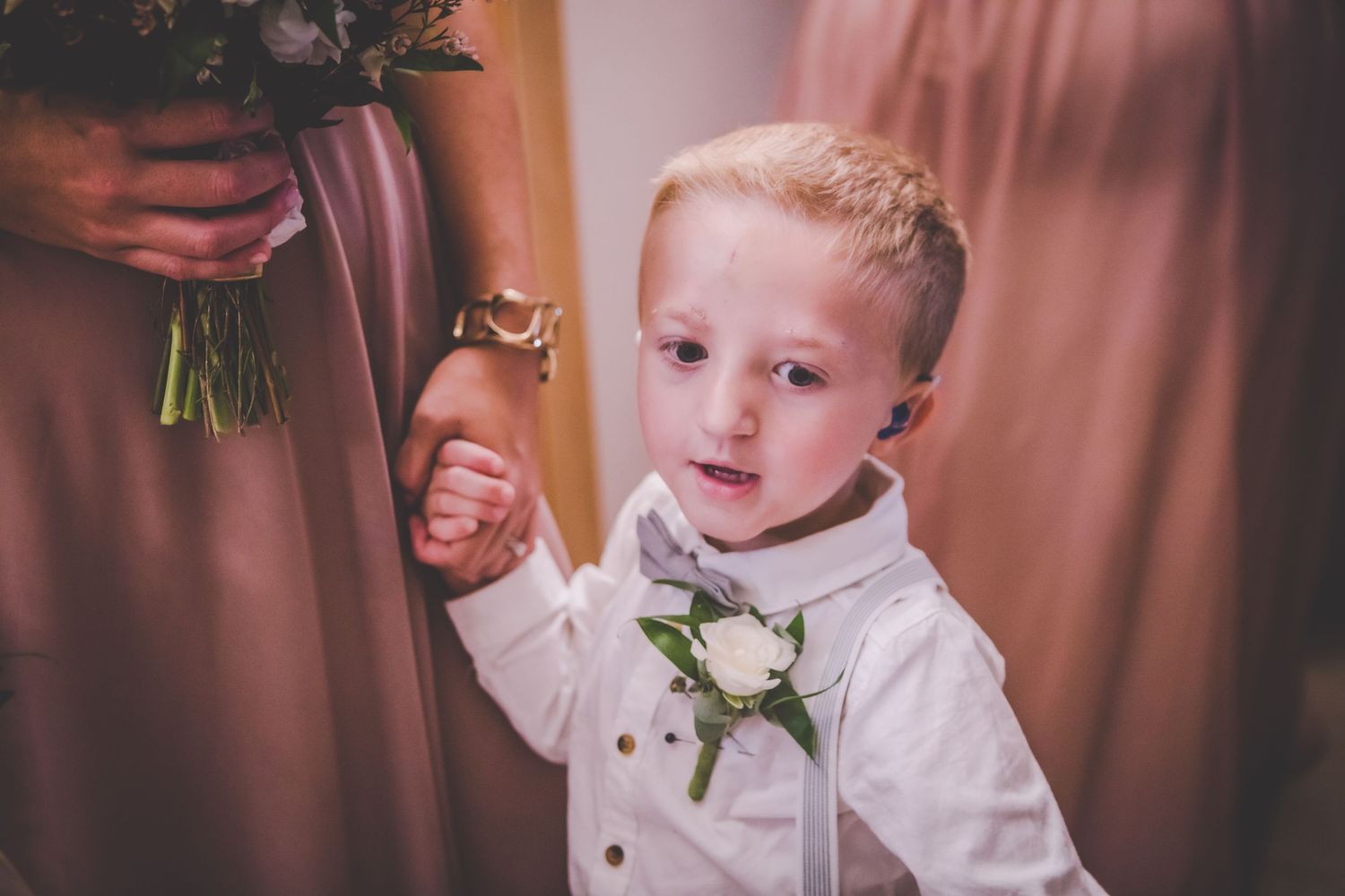 Young ring bearer in white suit holds hands with bridesmaid during wedding preparations.