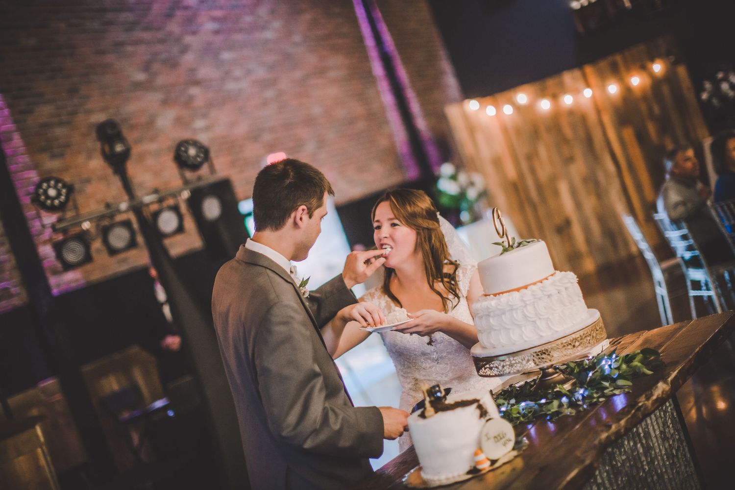 A couple laughs while sharing their wedding cake at a reception with exposed brick walls and twinkle lights.