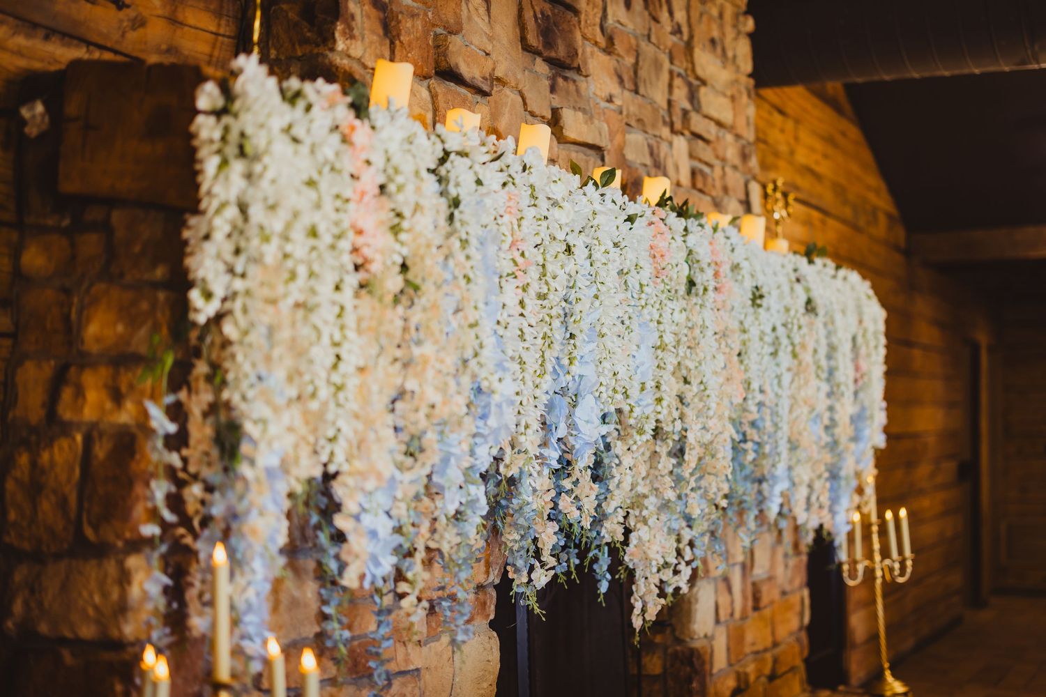 Elegant wedding backdrop with cascading white wisteria flowers and candles against a rustic stone wall.