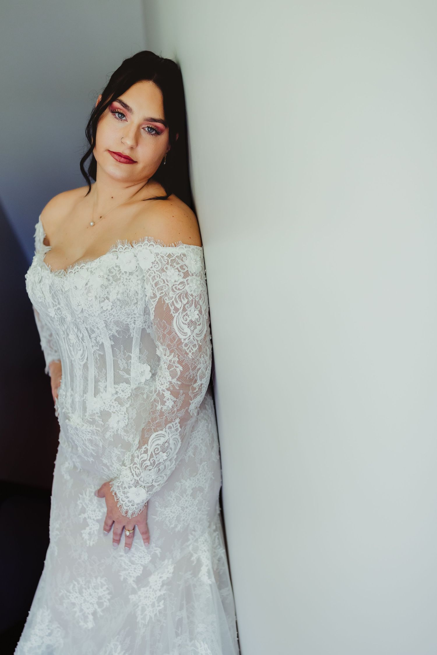 Bride in off-shoulder lace wedding gown with long sleeves leans against white wall, wearing bold red lipstick.