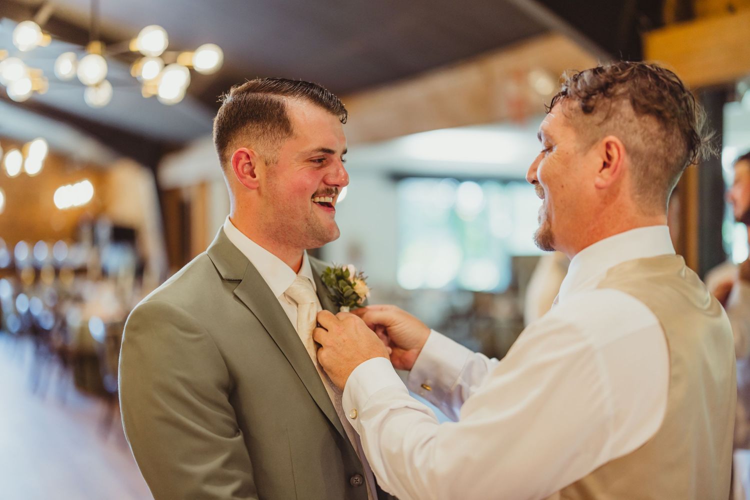 Groom laughing as man pins boutonniere on his gray suit lapel at wedding reception.