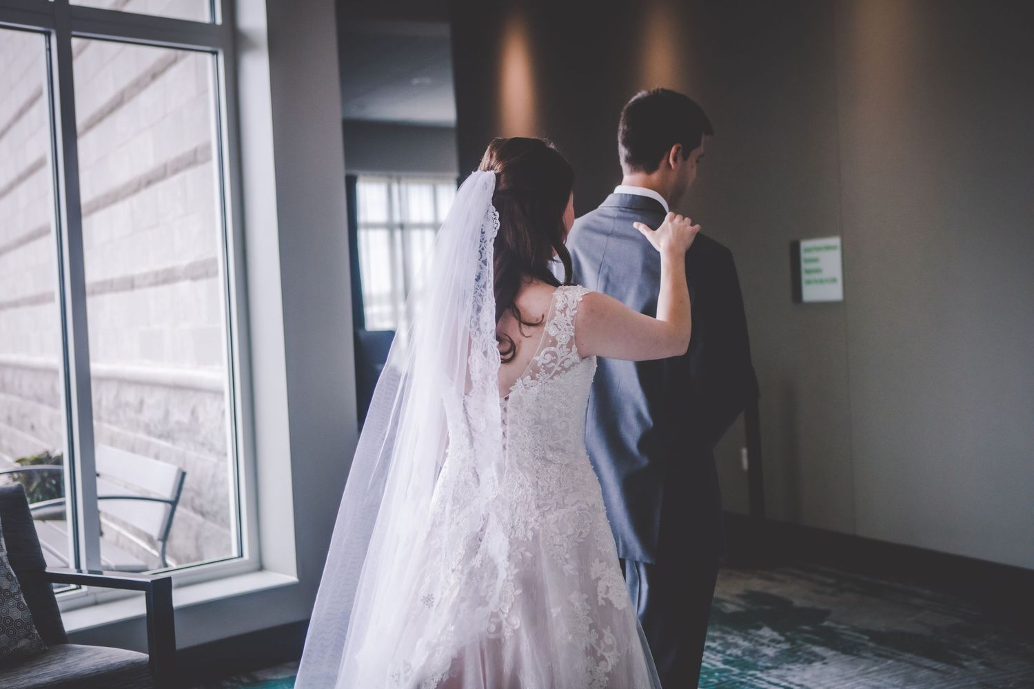 Candid first look moment between newlyweds by large windows in modern venue.
