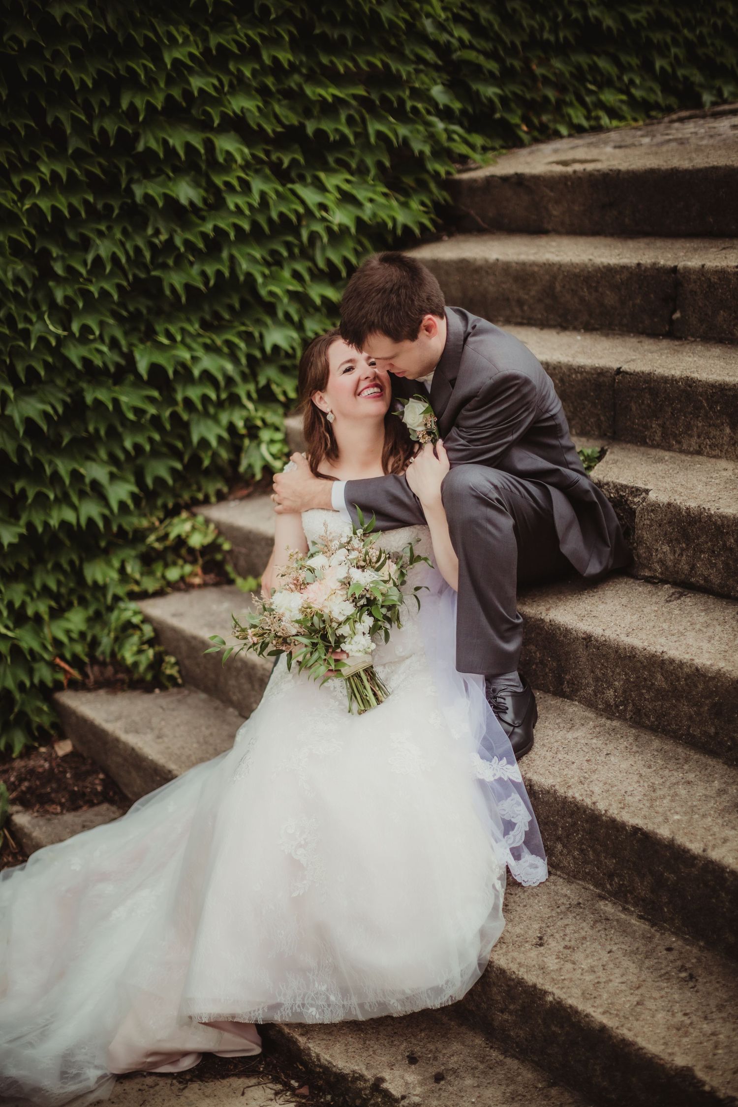 A romantic wedding photo of a couple laughing together on ivy-covered stone steps outdoors.