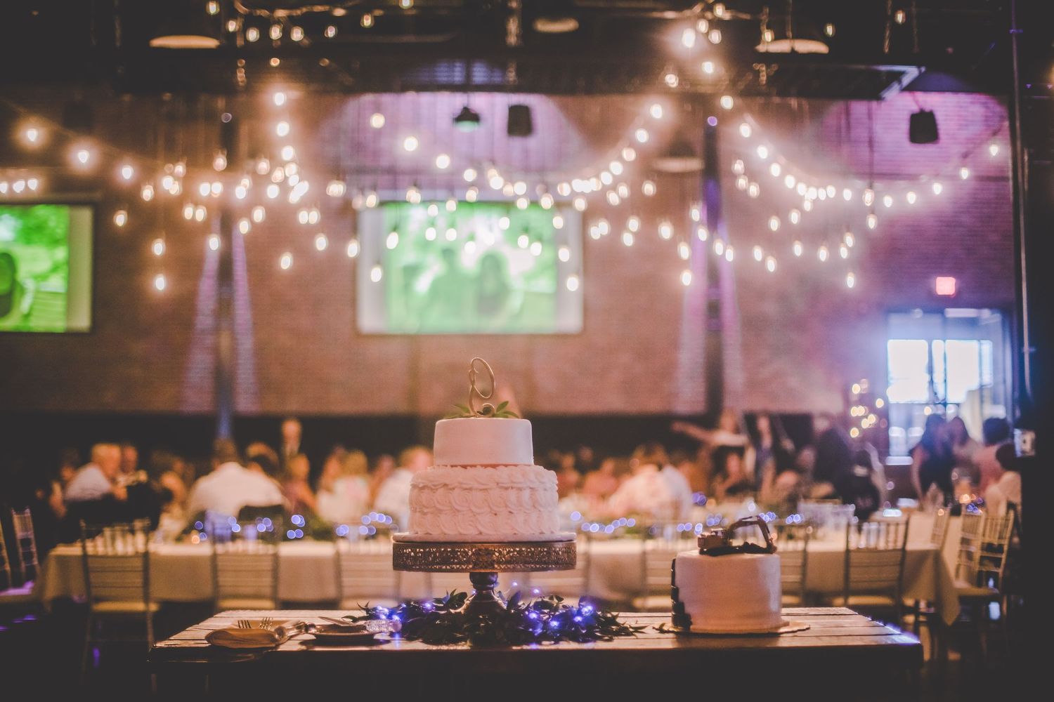 Wedding reception venue with string lights, screens, and tiered cake centerpiece.