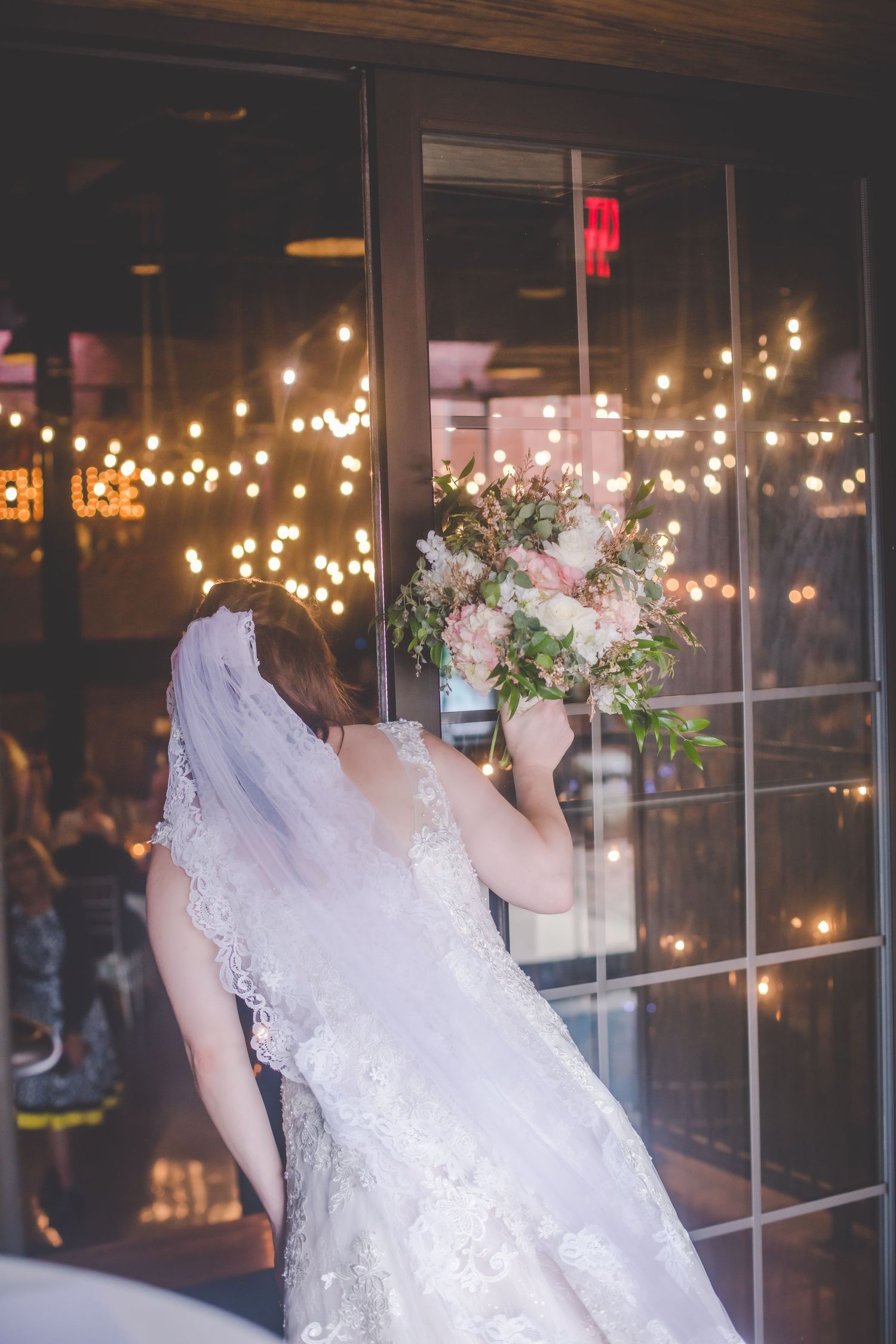 A bride holding a floral bouquet near a window with string lights in the background.