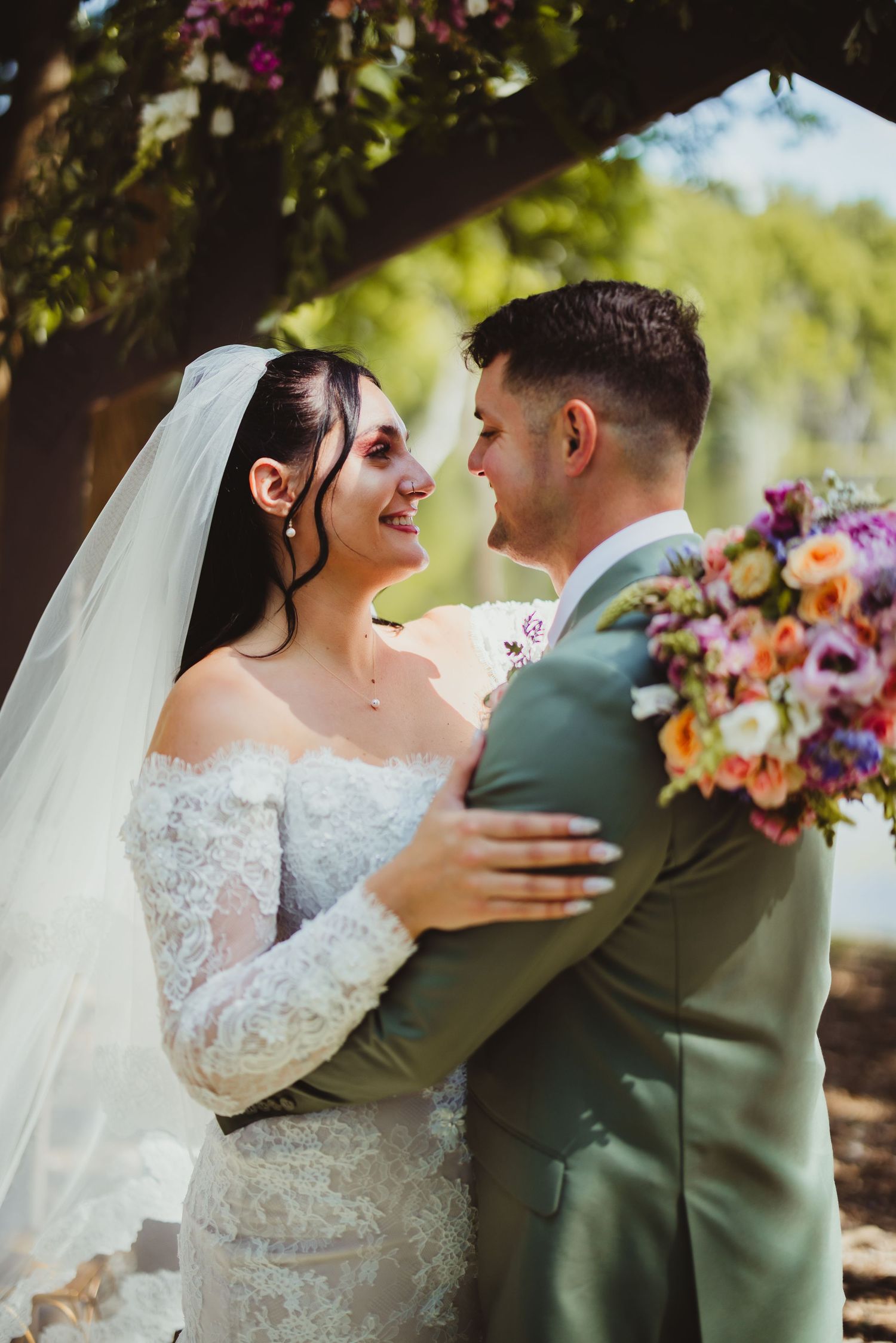 Bride in lace off-shoulder gown and veil smiles at groom in sage suit holding colorful bouquet outdoors.