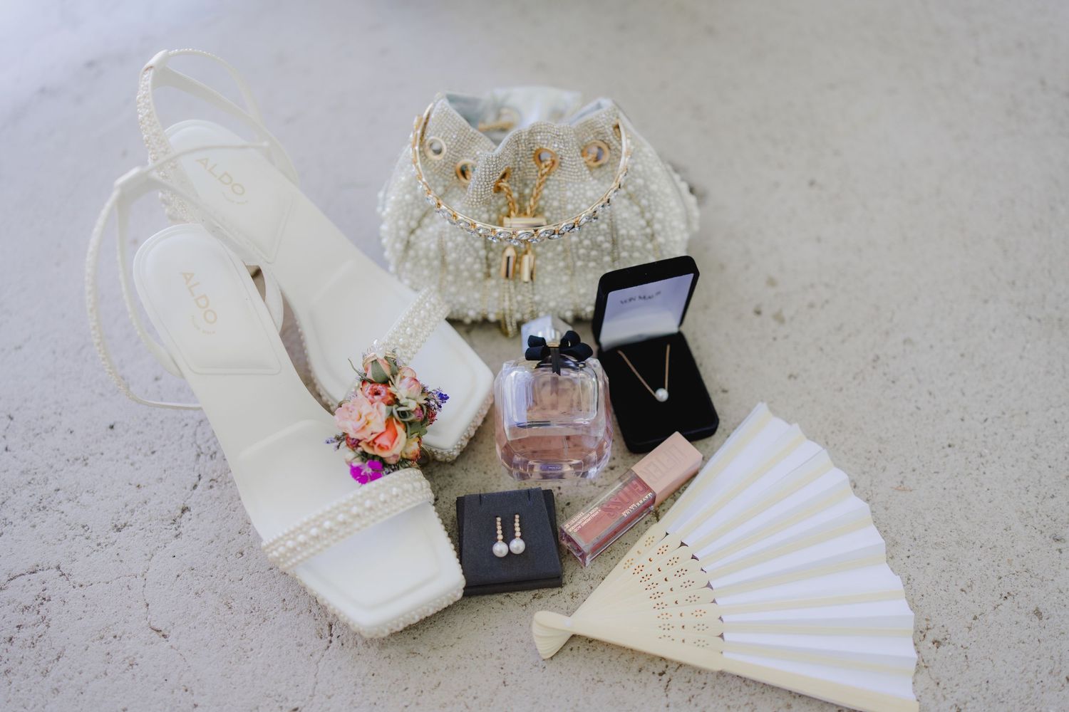 White bridal heels, pearl clutch, perfume, necklace, earrings, lipgloss, and a white fan arranged elegantly on a surface.