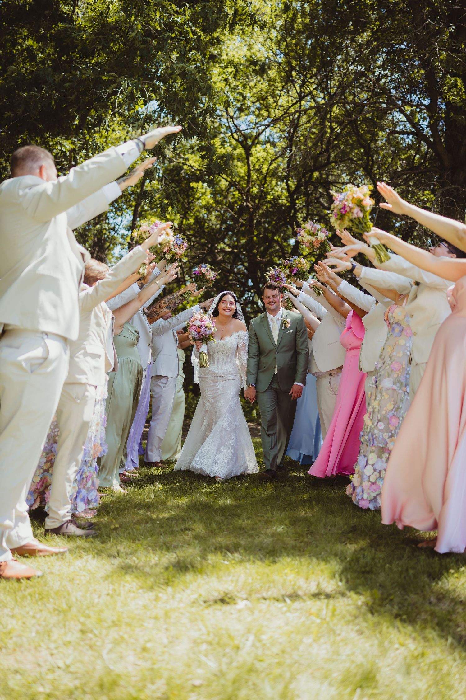 Bride and groom walk through an arch of raised bouquets held by wedding party in pastel attire outdoors.