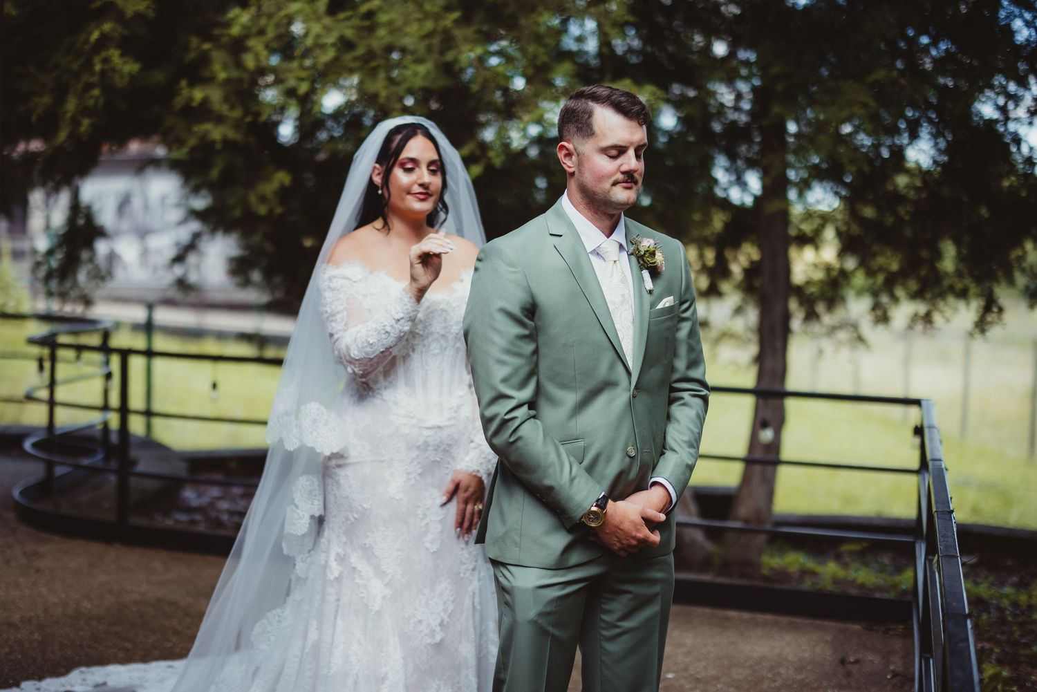 Bride in lace gown and veil stands beside groom in sage suit outdoors near a railing surrounded by trees.