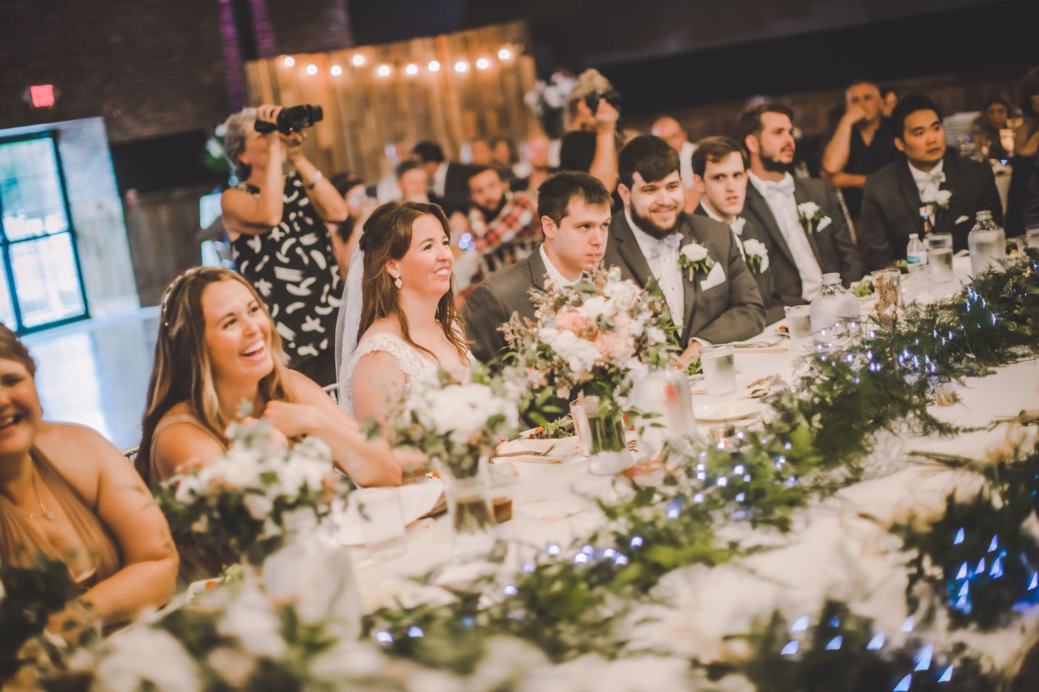 Wedding party and guests seated at a long decorated table with floral centerpieces and string lights overhead.