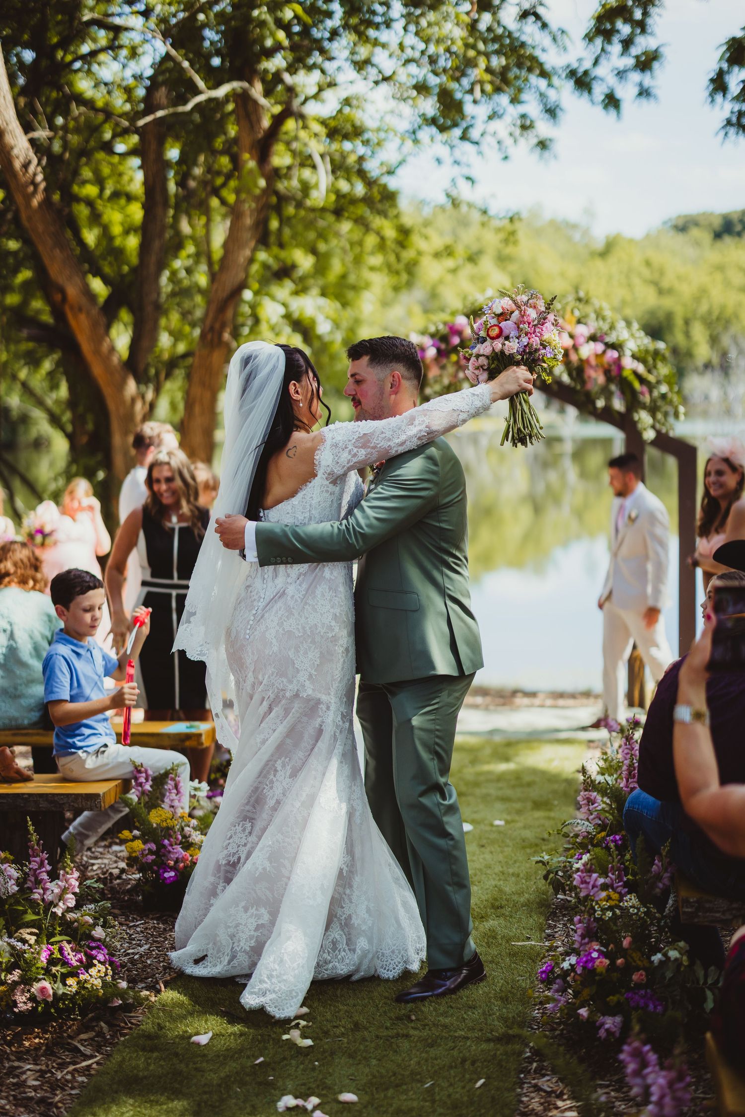 Bride and groom share first kiss at outdoor lakeside wedding ceremony, surrounded by purple floral arrangements and guests.