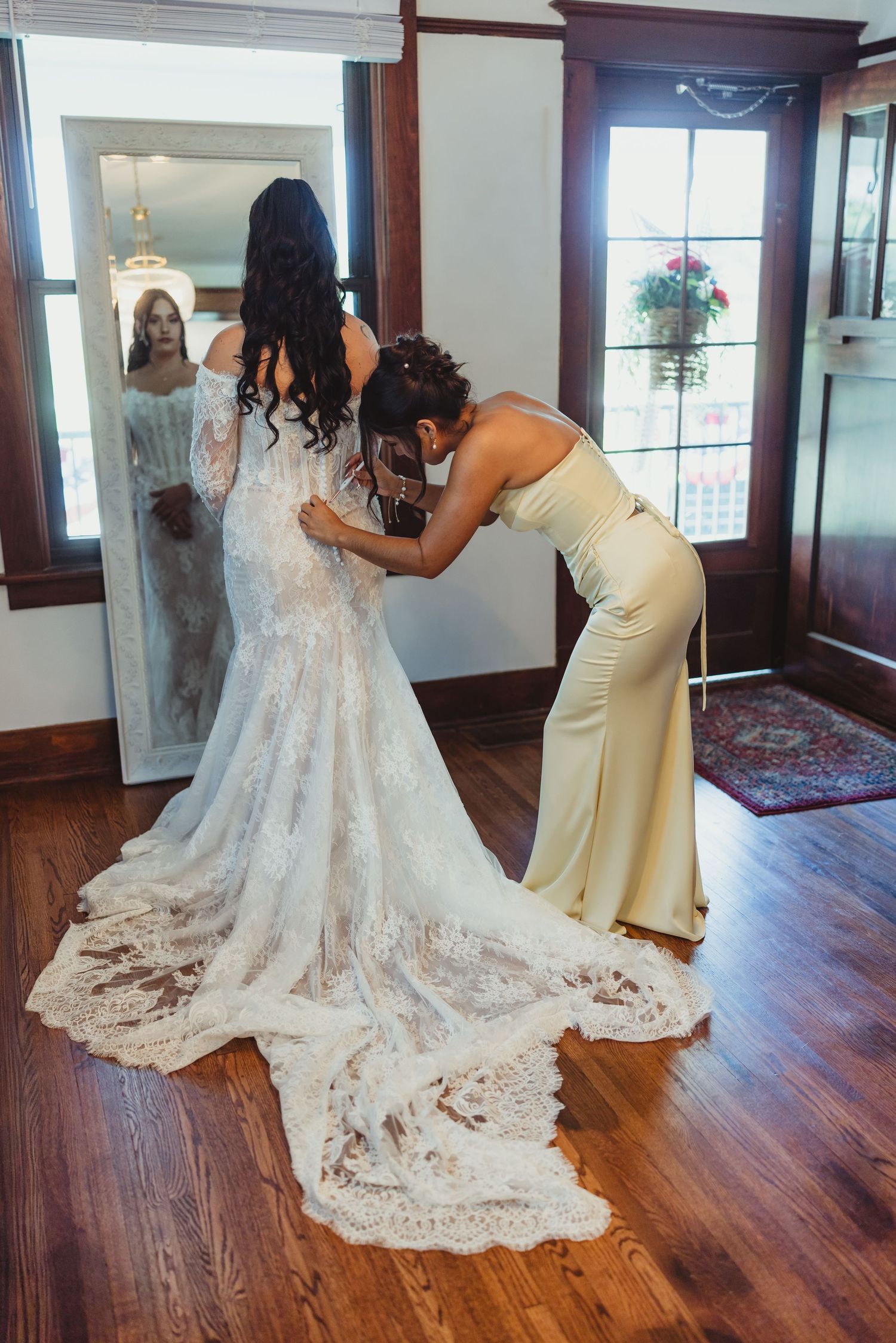 Bridesmaid in gold dress fastening the back of a bride's lace wedding gown with a long train indoors.