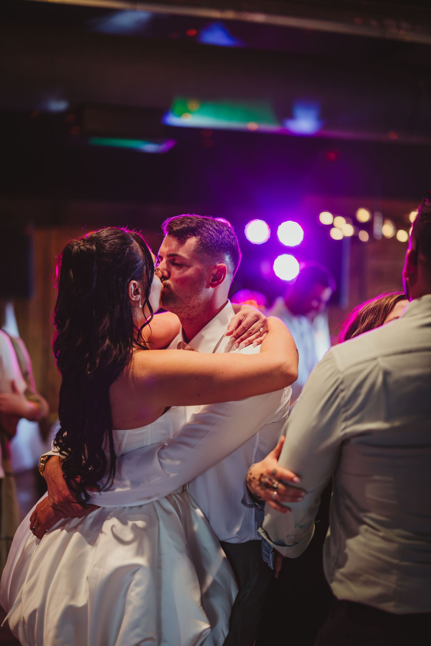 Bride and groom sharing a kiss on the dance floor surrounded by guests at wedding reception.