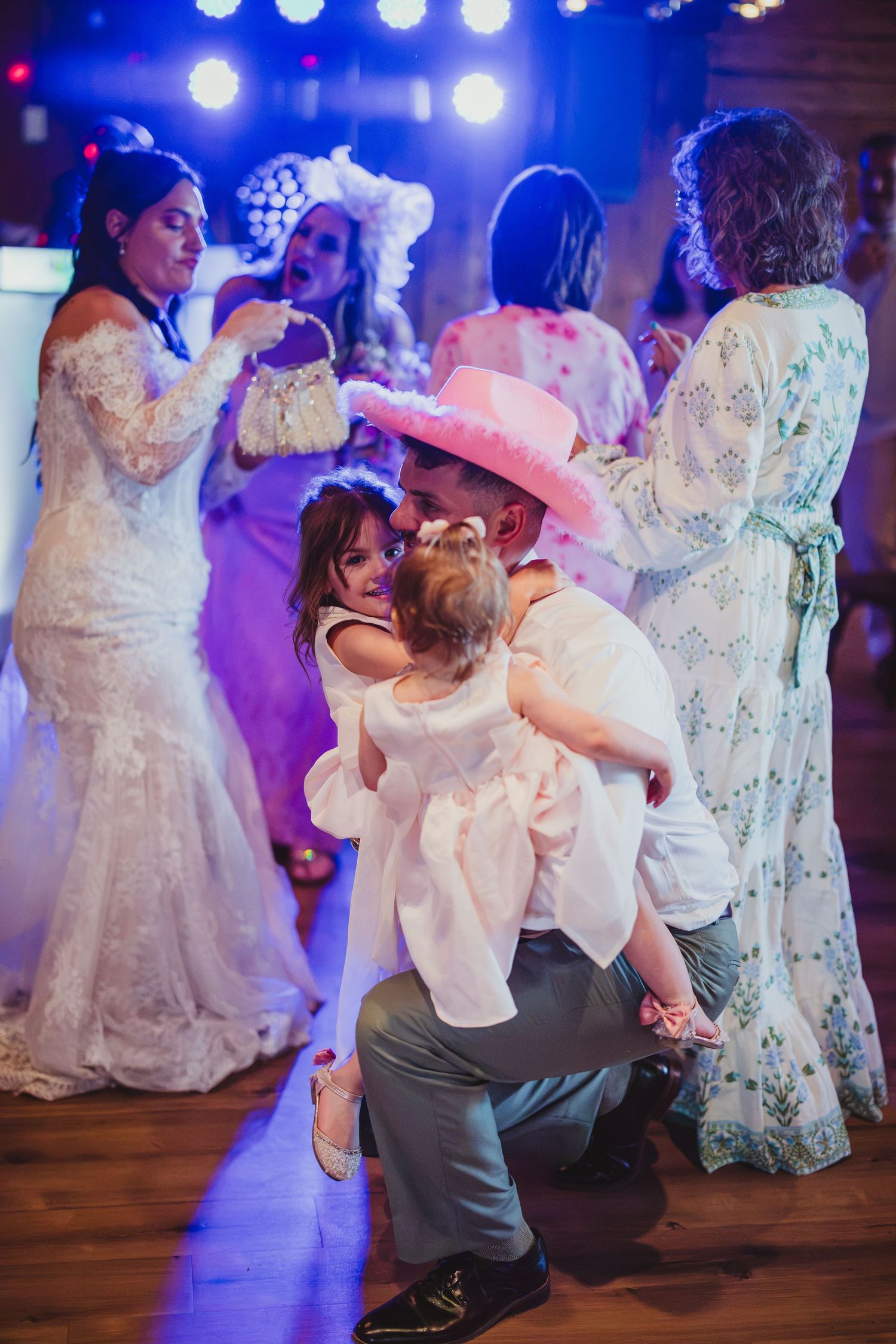 Man in pink cowboy hat kneeling to embrace young girl on wedding dance floor with guests dancing.
