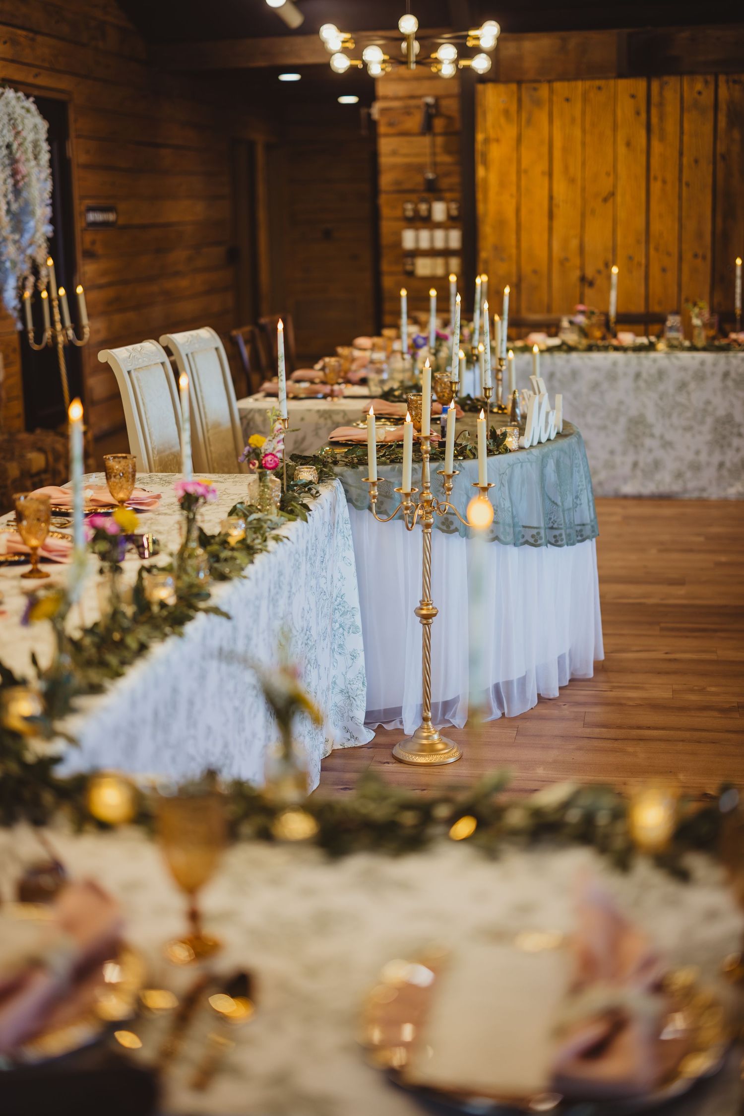 Romantic barn wedding head table with white lace runner, gold candelabras, greenery garland, and glowing candles.