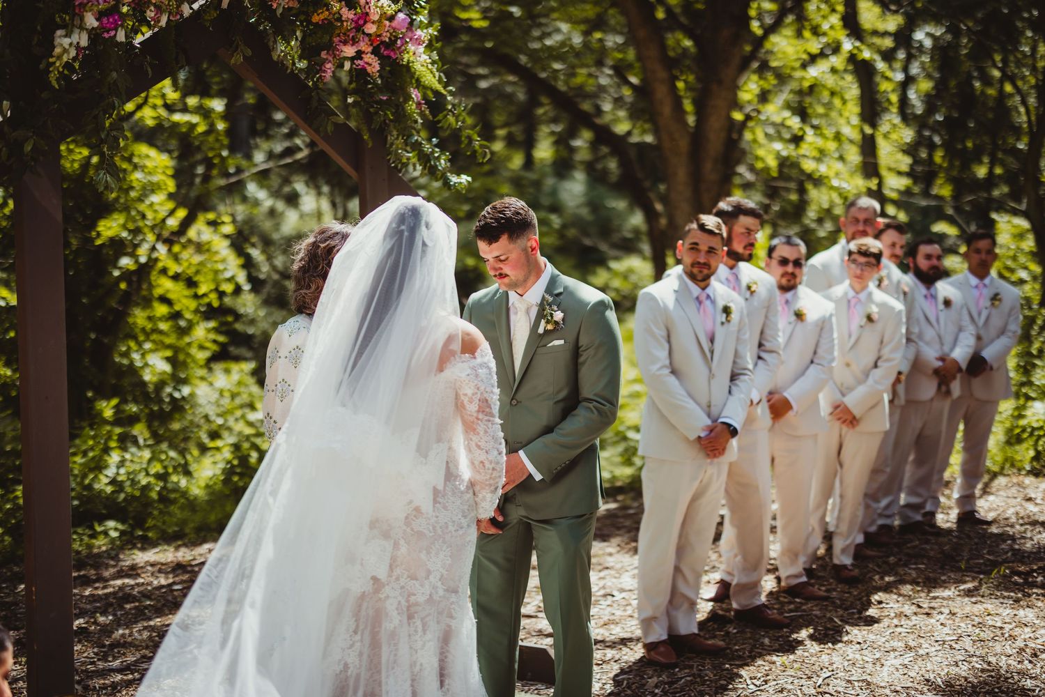 Groom in sage green suit faces bride in lace wedding dress at outdoor ceremony with groomsmen in tan suits.