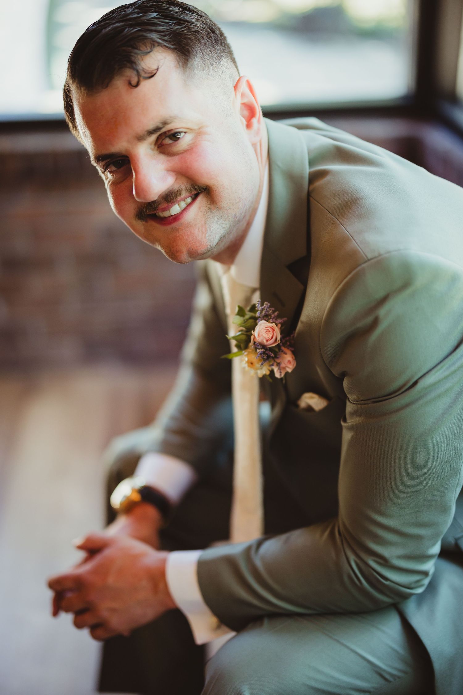 Smiling groom in sage green suit with floral boutonniere poses seated, leaning forward with hands clasped at wedding.
