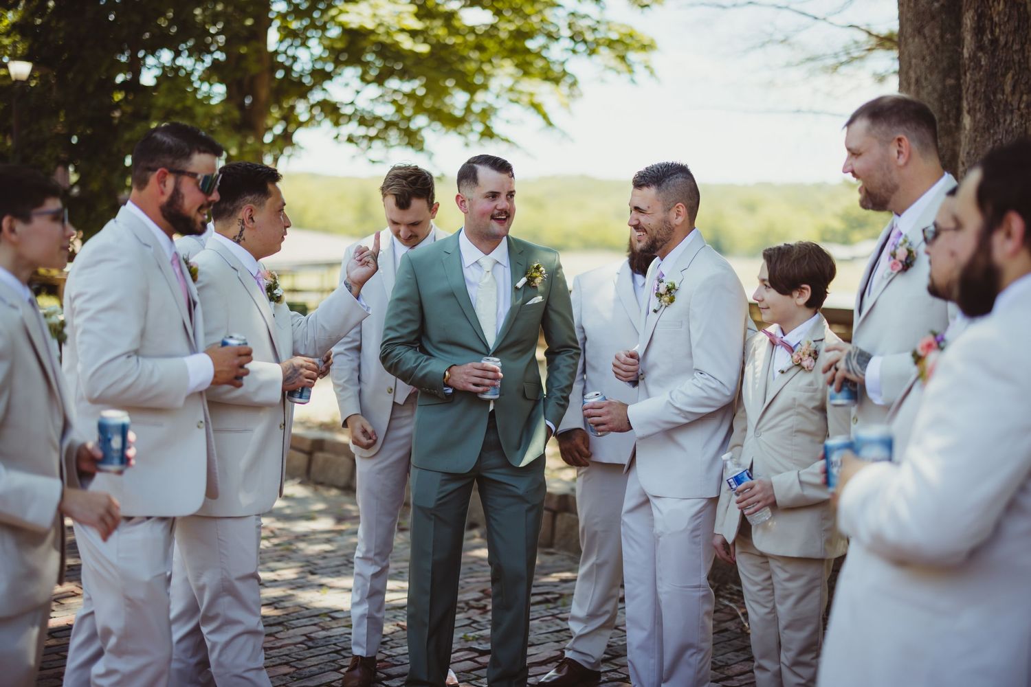 Groom in green suit stands with groomsmen in tan suits, laughing outdoors at a rustic wedding celebration.