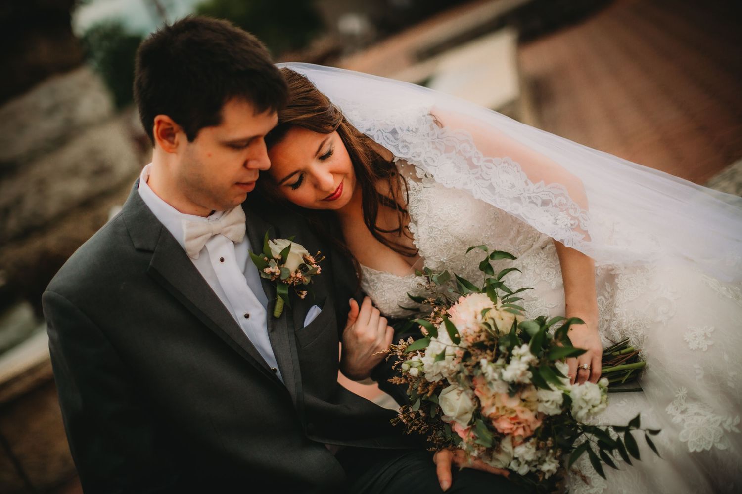 Wedding couple shares intimate moment with bridal bouquet of white and peach flowers.