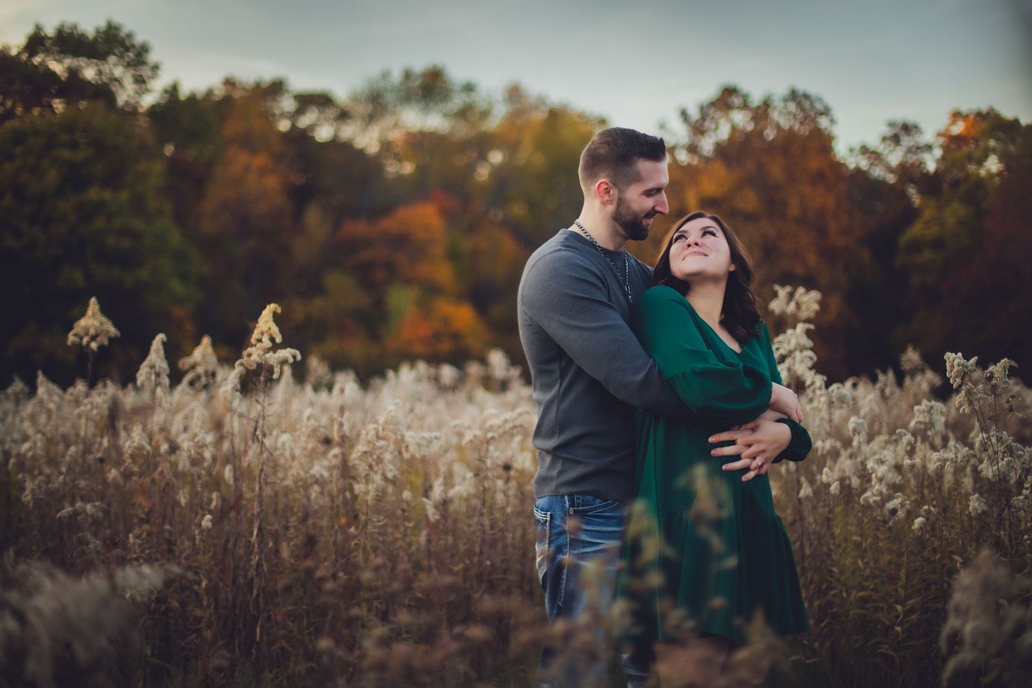 A couple shares an intimate embrace surrounded by autumn scenery and tall grass.