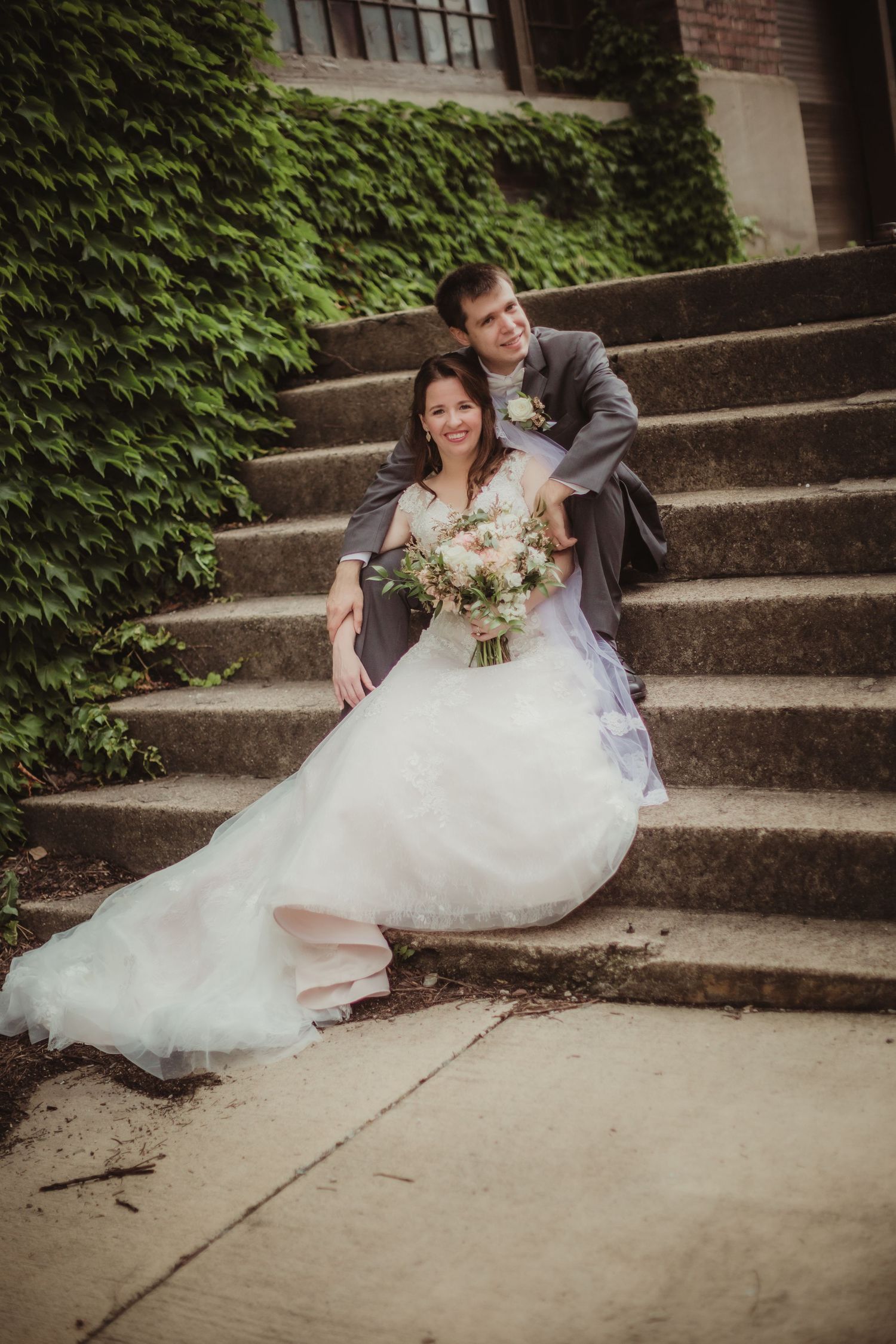 A wedding couple relaxes on stone steps with lush green ivy backdrop during their photoshoot.