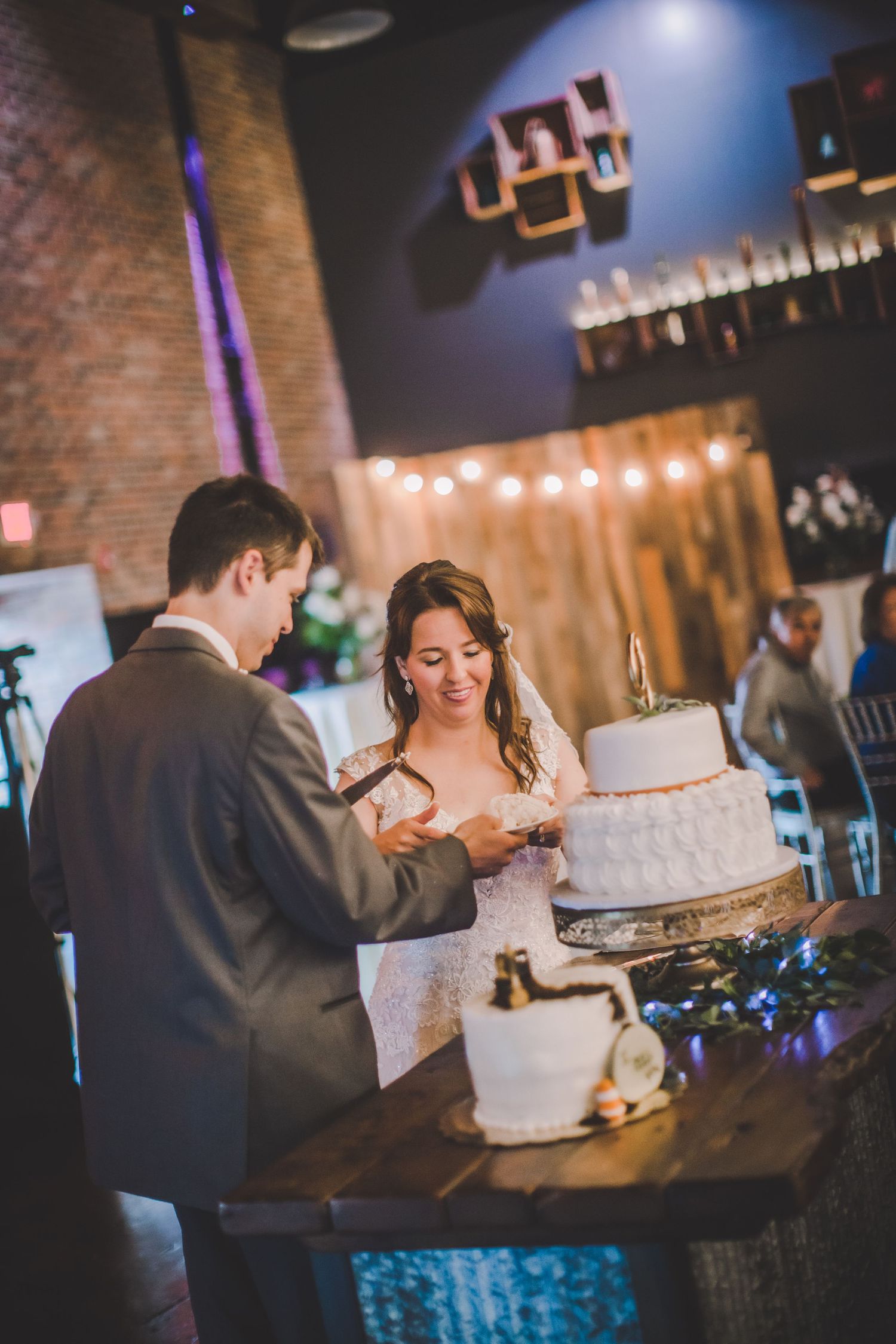 A couple sharing their first slice of wedding cake together at their wedding reception with string lights and brick walls.