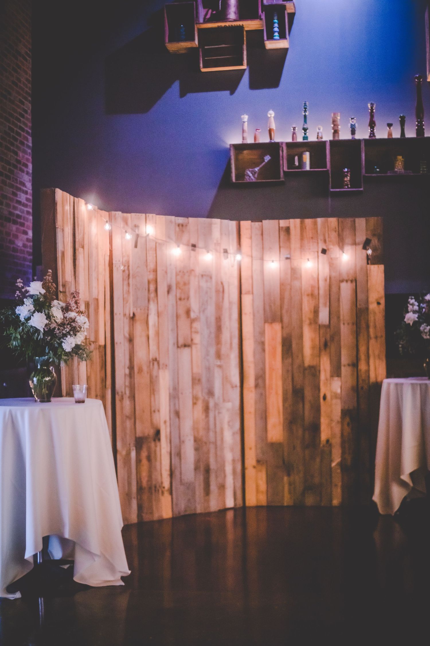 Rustic wedding venue setup with wooden backdrop, string lights, and bottle display shelves.