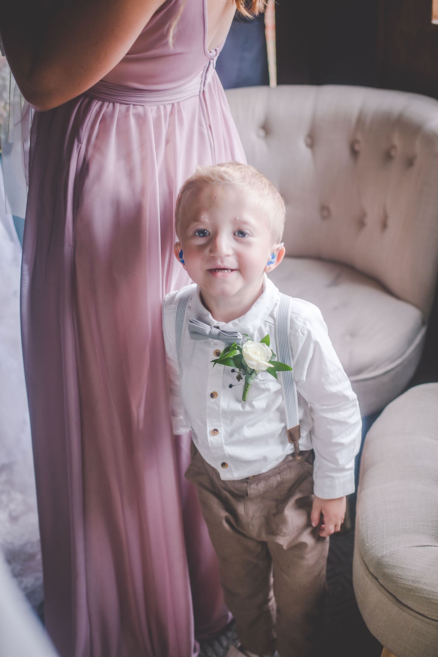 A small ring bearer in suspenders and bowtie stands by a grey couch.