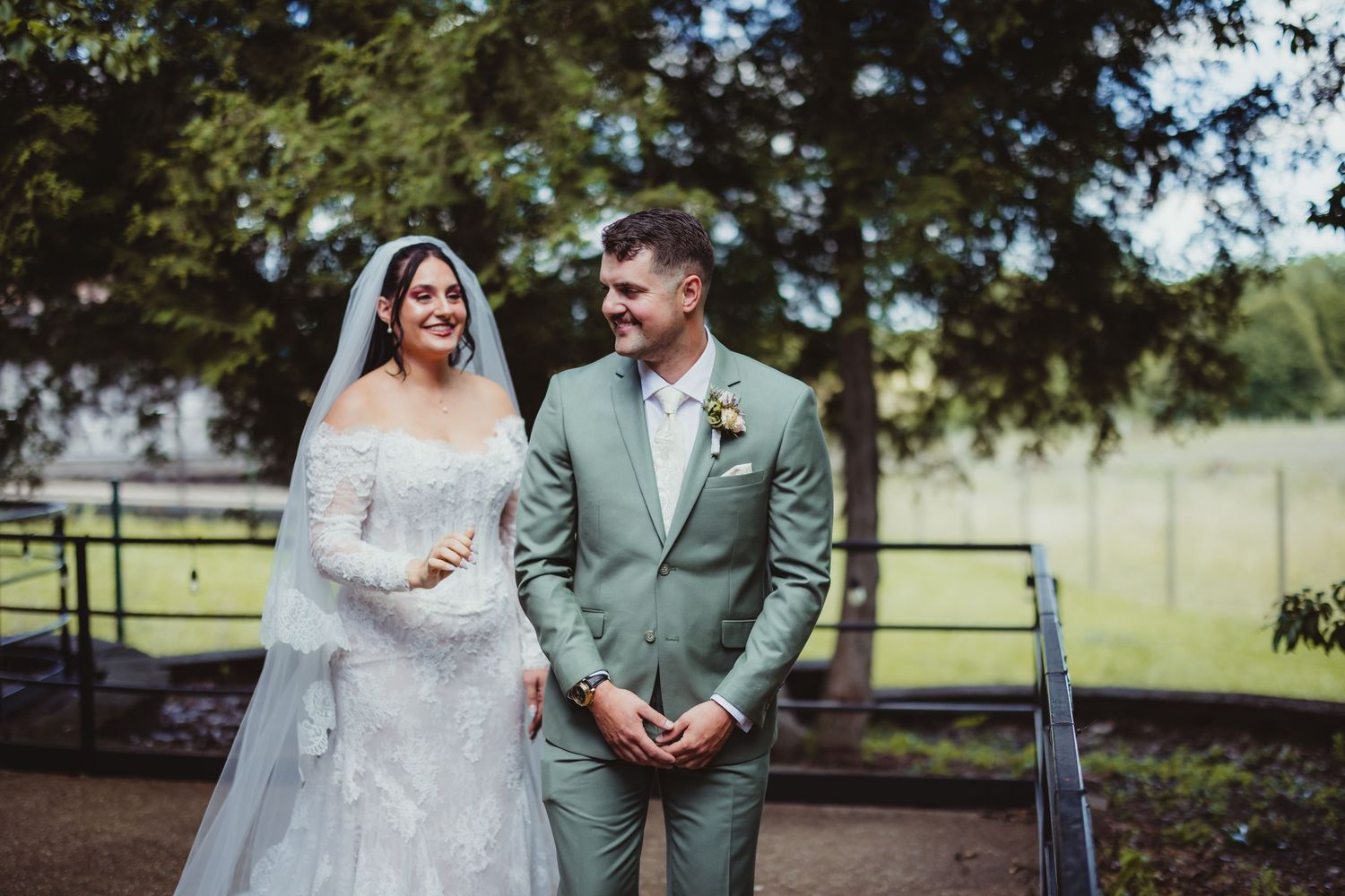 A bride in a white lace gown and groom in a sage green suit share a romantic moment outdoors.