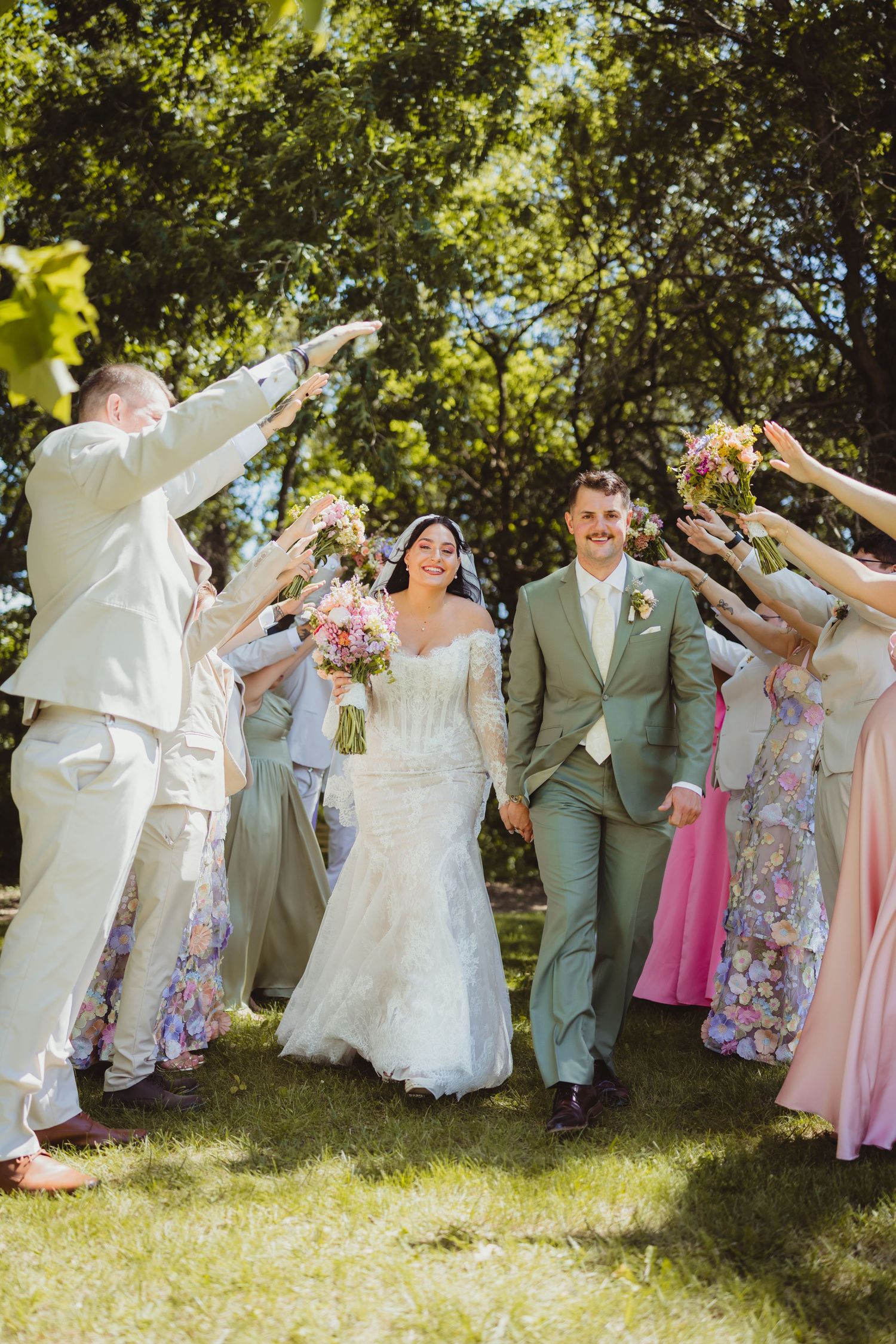 Bride and groom walk through a celebratory arch of raised hands outdoors, surrounded by wedding party in sage and pink attire.