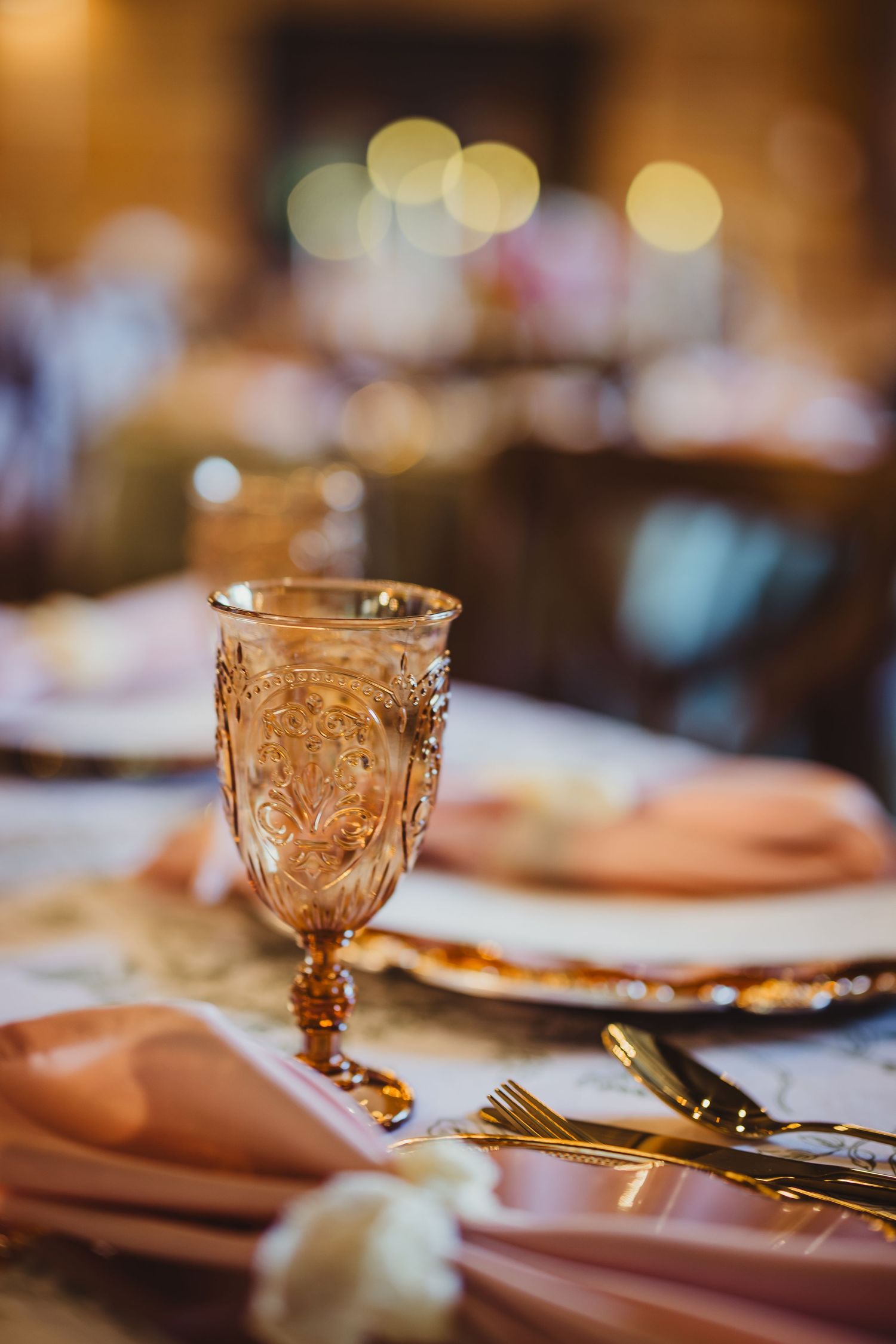 Elegant wedding table setting with ornate pink goblet, gold cutlery, blush napkins, and decorative plates in warm bokeh lighting.