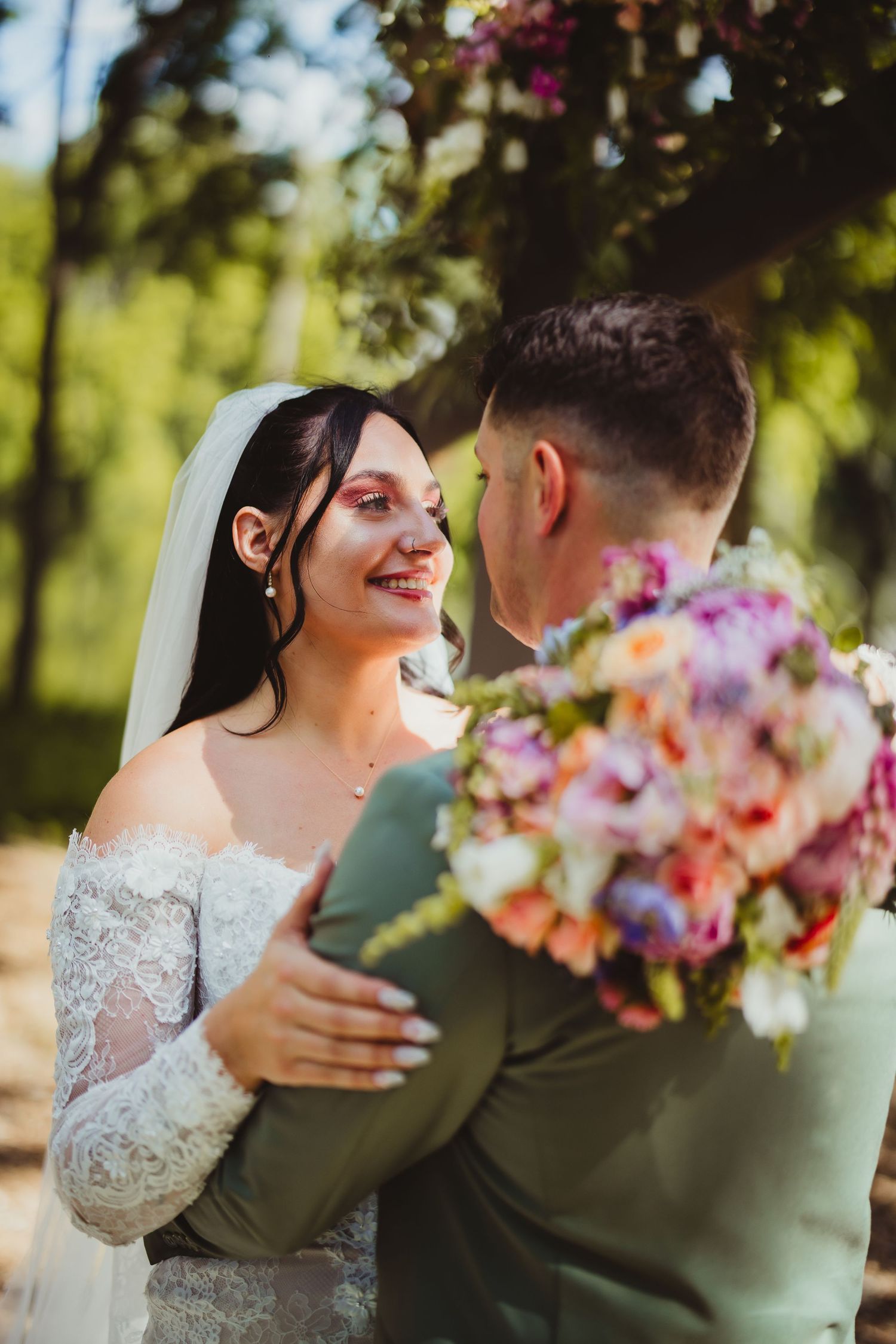 Smiling bride in lace gown embraces groom outdoors under floral arch holding colorful bouquet.