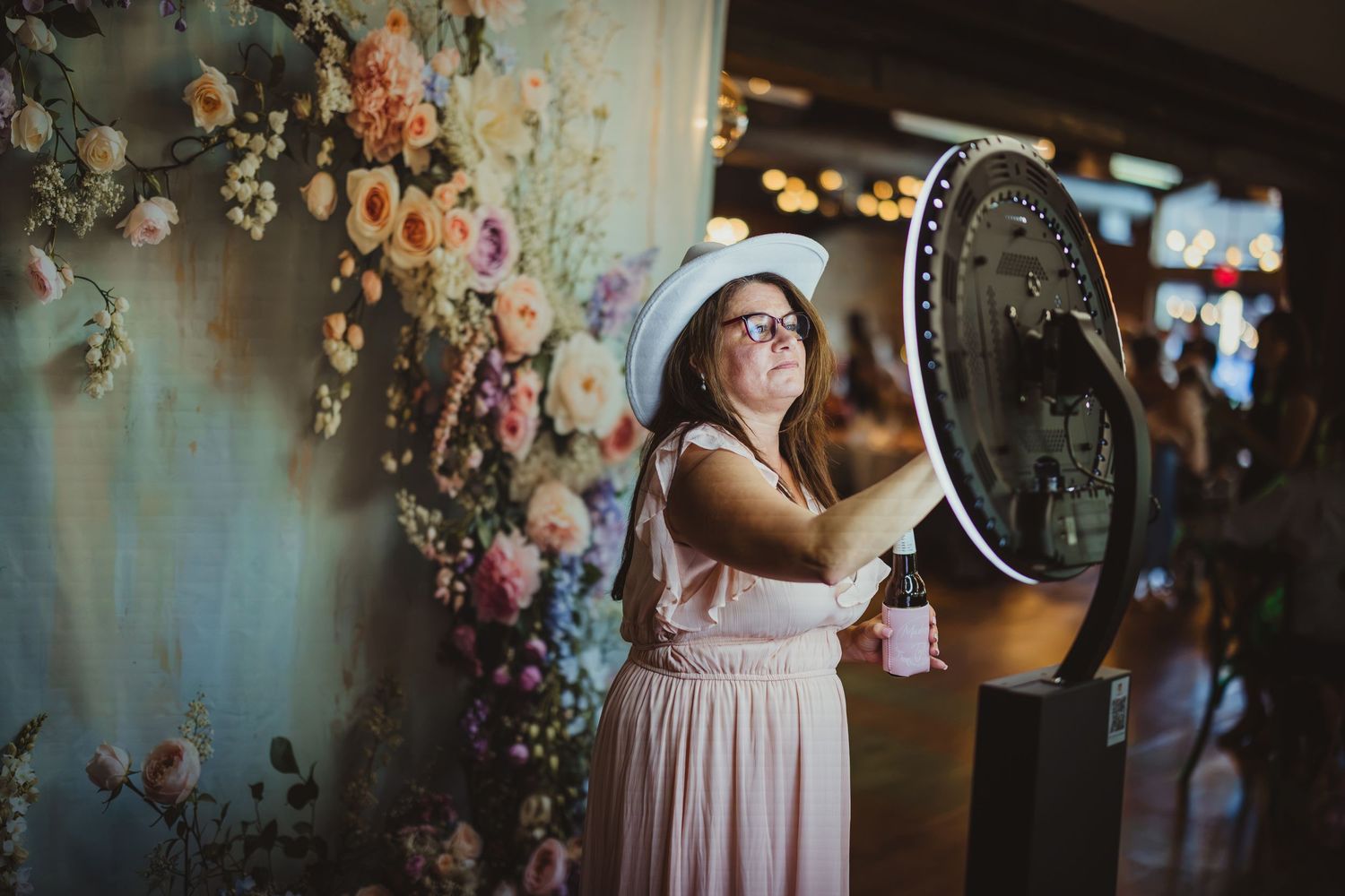 Woman in pink dress and white hat poses with a circular photo booth mirror against a floral backdrop.