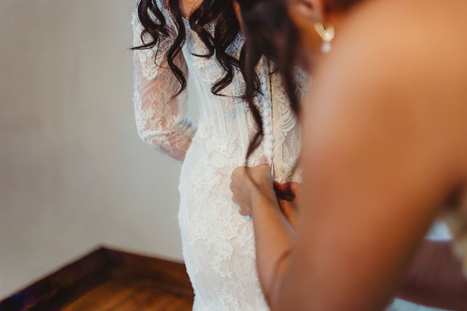 Bride in lace wedding gown being helped into dress by bridesmaid, elegant bridal preparation moment.