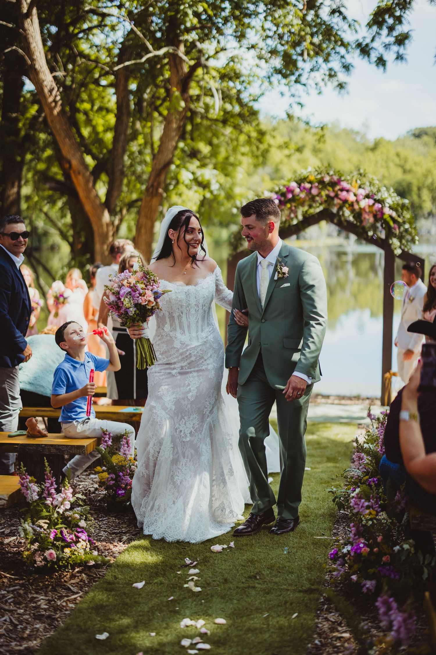 Newlyweds walk joyfully down the aisle after outdoor lakeside wedding ceremony surrounded by guests.