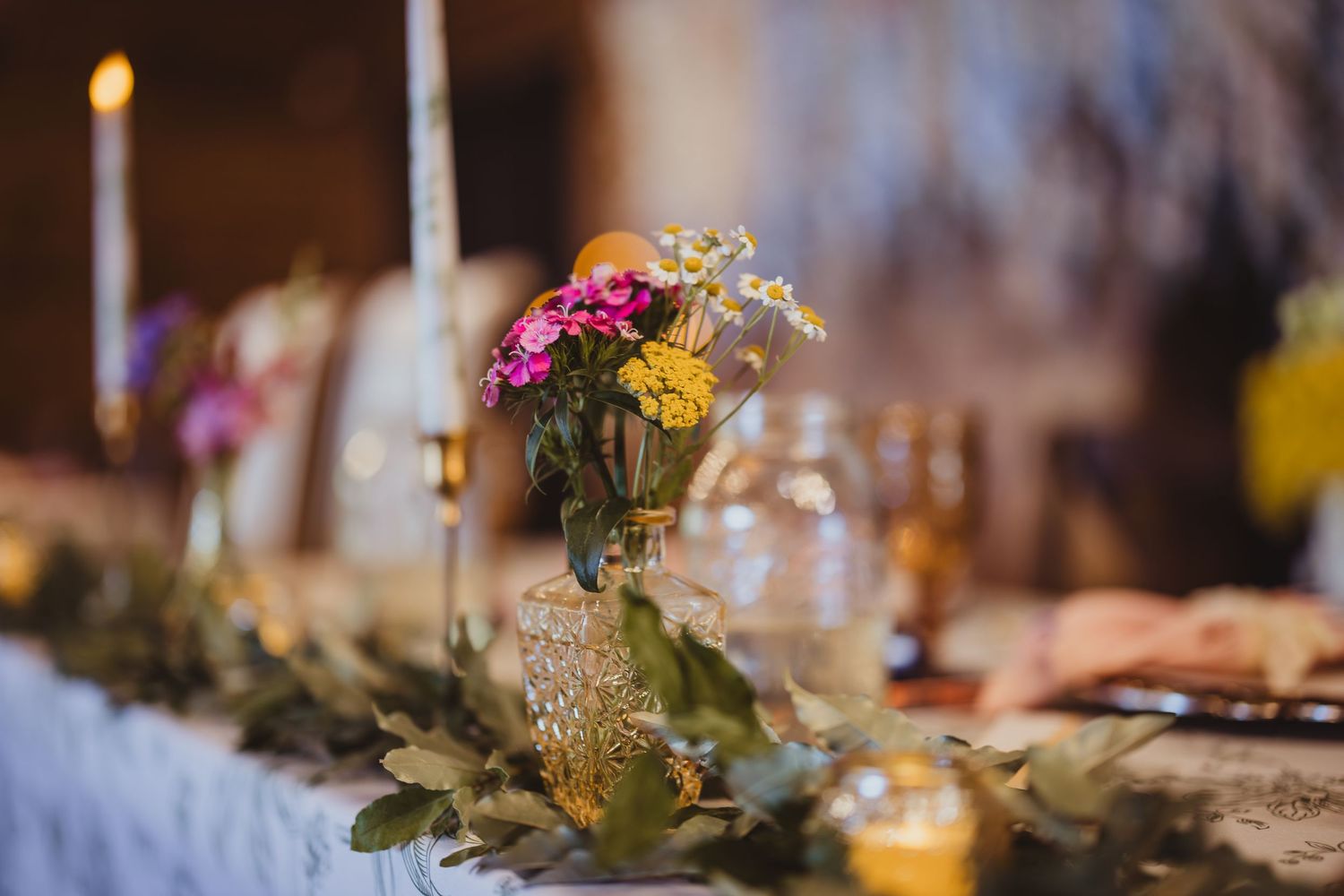 Elegant wedding table centerpiece with pink and yellow flowers in gold vase, greenery garland, and candles.